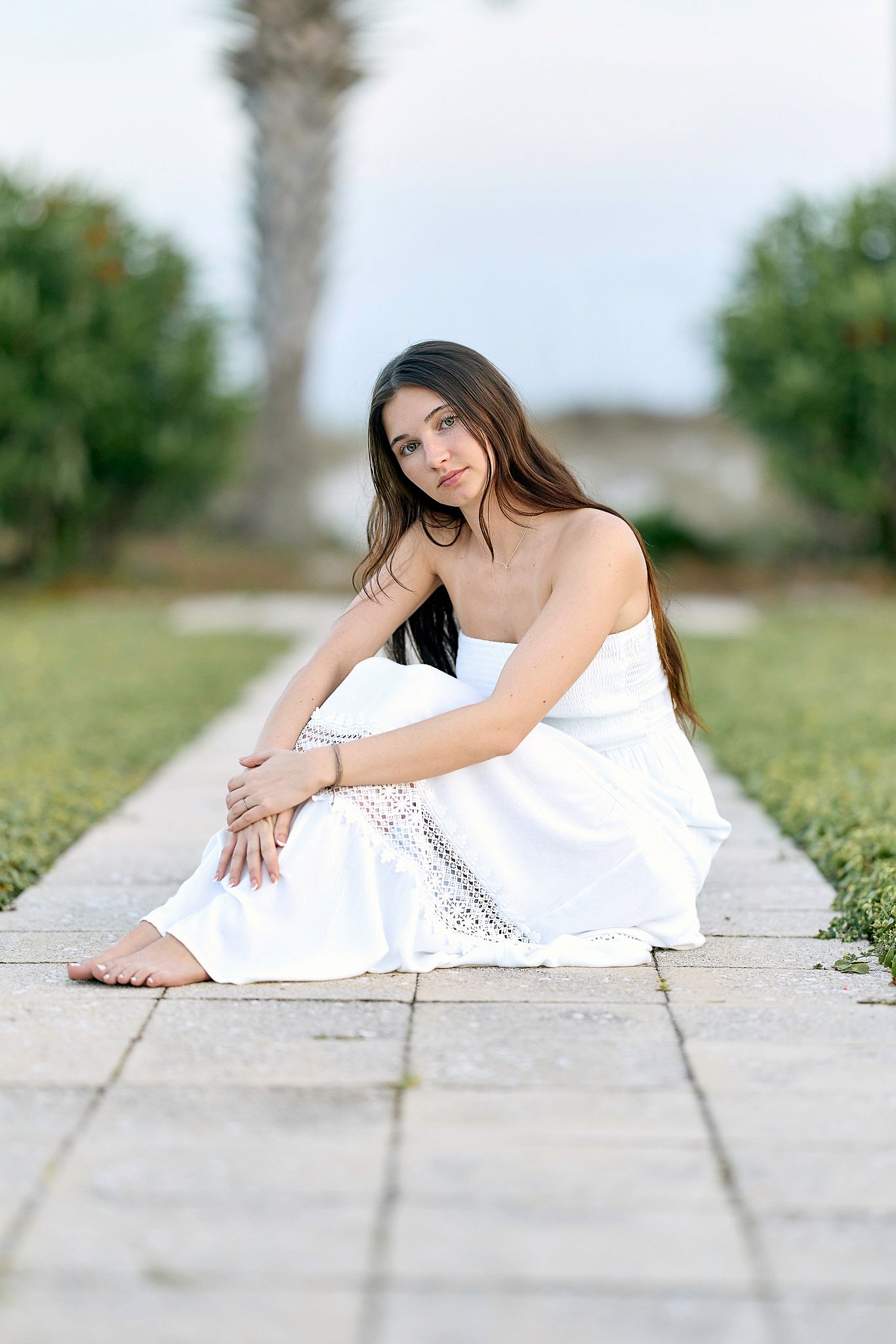 Someone in a flowing white dress sits gracefully on stone pavers with palm trees and greenery in the background.