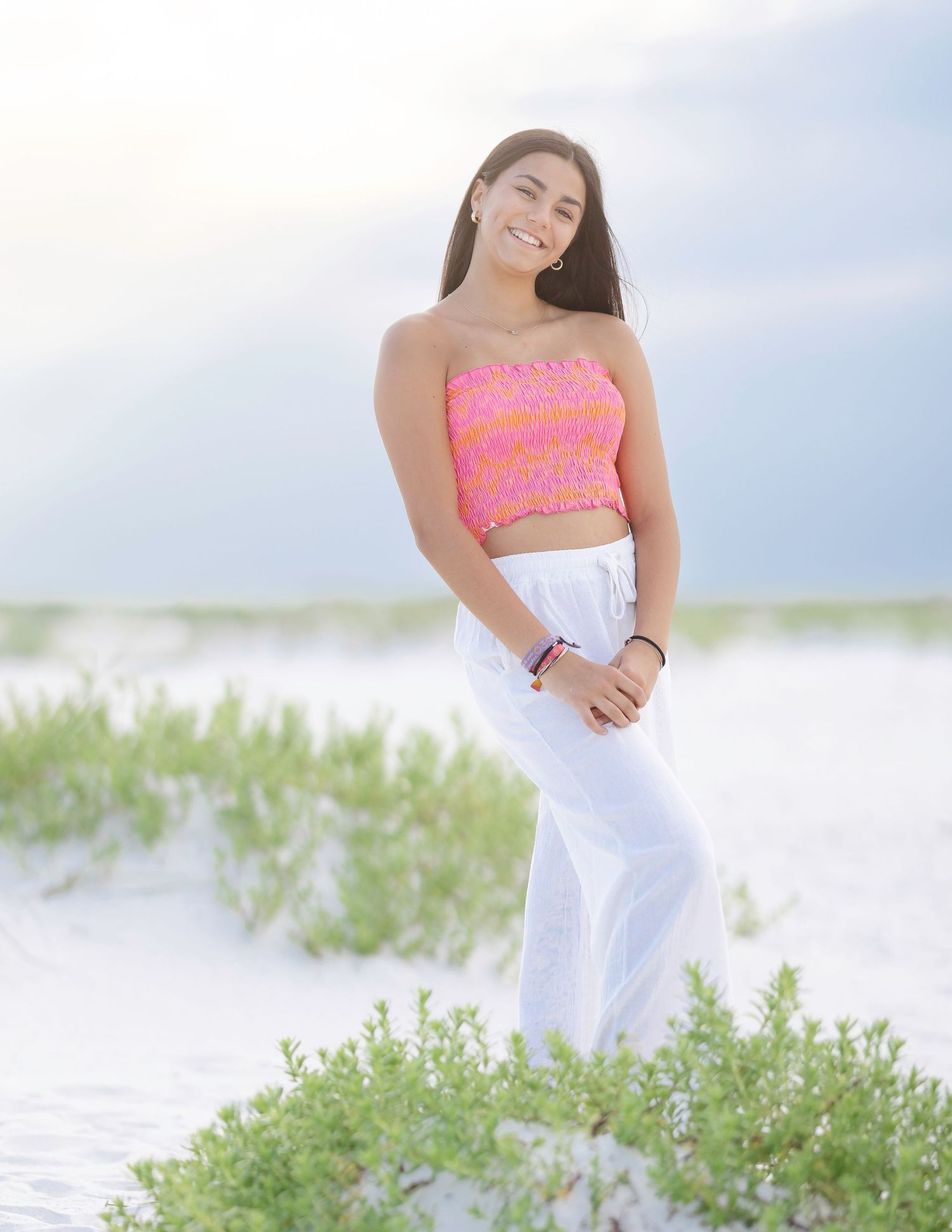A person in a pink top and white skirt poses gracefully in a field of green coastal vegetation near the beach.