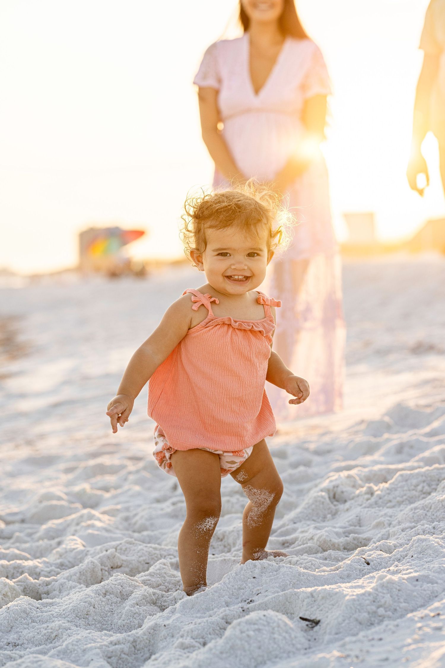 Child plays and jumps joyfully on a sandy beach at sunset in a coral-colored romper.