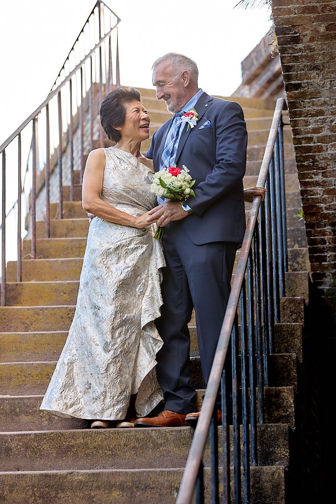 A joyful couple shares a laugh on stone steps during their wedding, with the bride holding a pink and white bouquet.