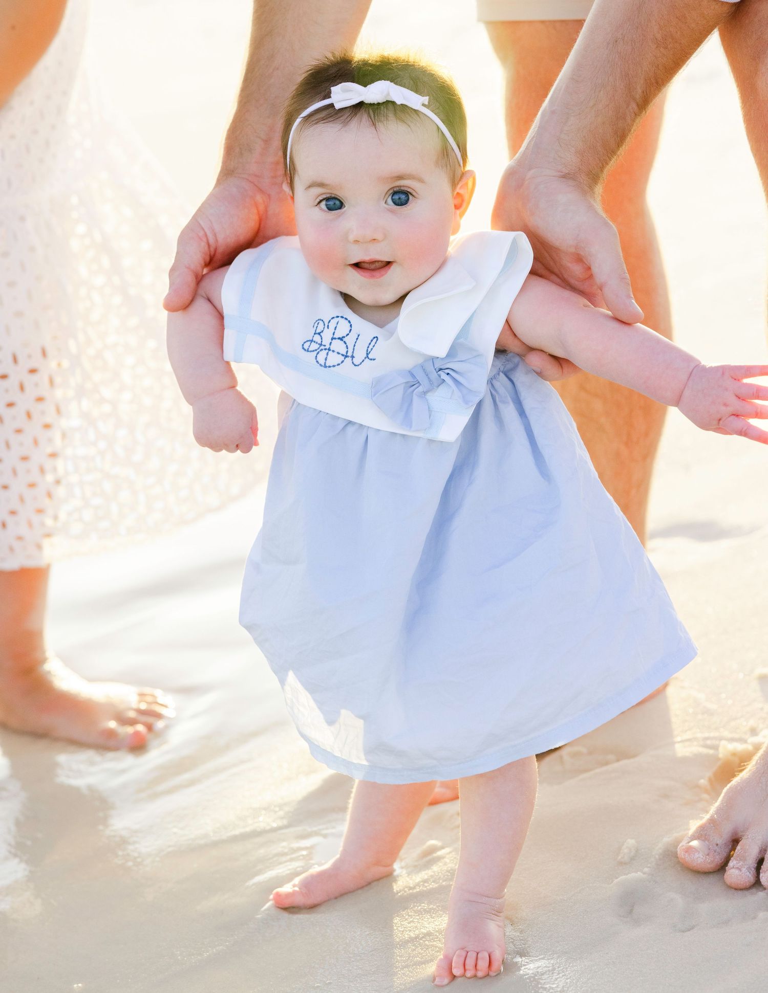 Baby in light blue dress crawling on white sand at the beach.