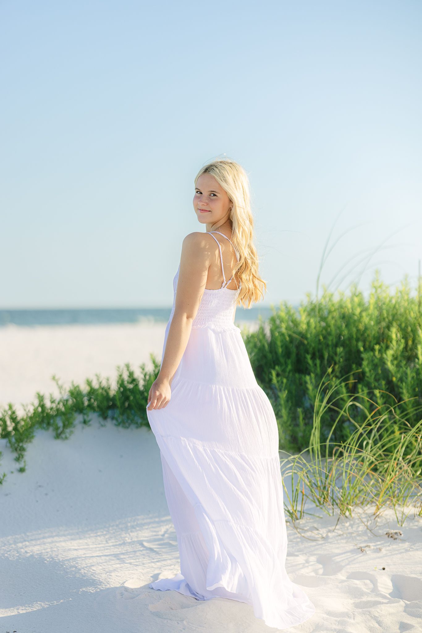 A person wearing a white sundress poses gracefully among beach grass and sand dunes.