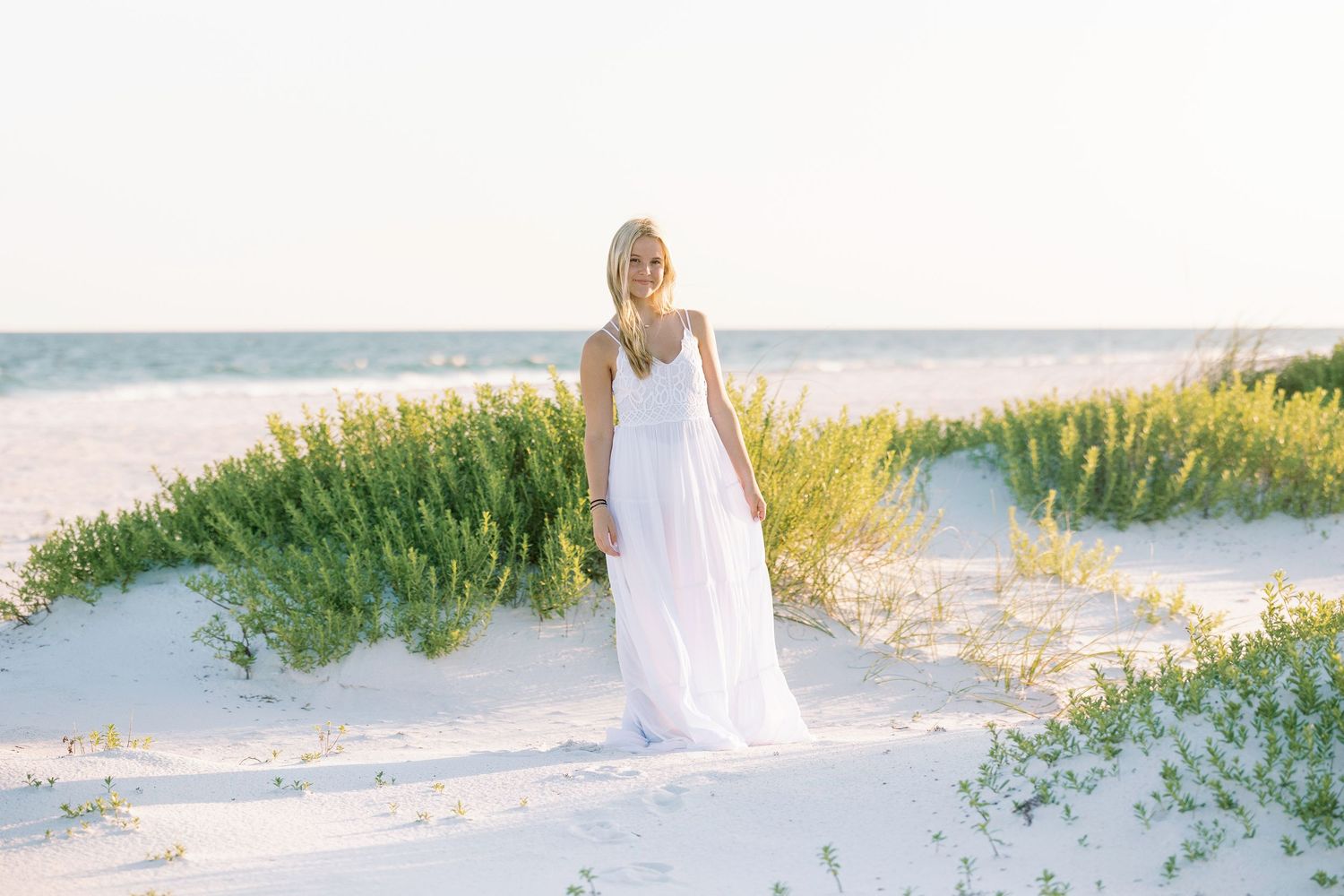 Person in flowing white dress stands among dunes and sea oats on pristine beach.