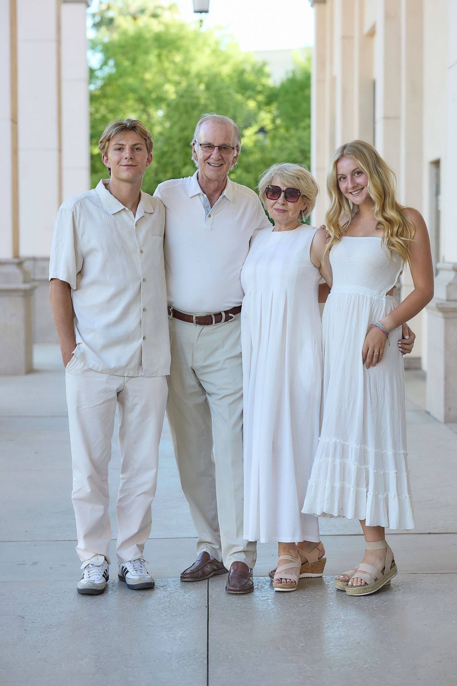 Four people wearing coordinated white outfits pose together on a covered walkway with columns in the background.