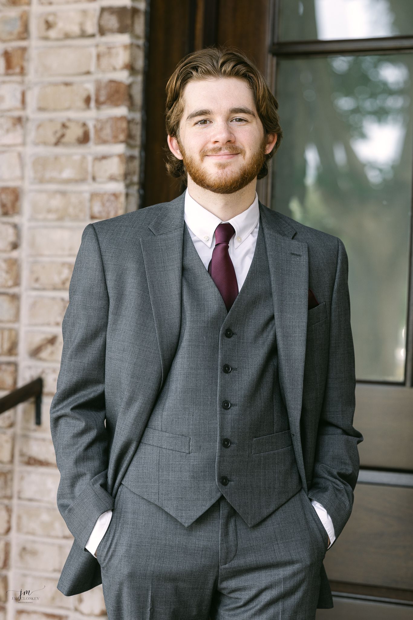 A man in a grey three-piece suit and burgundy tie stands confidently against brick architecture.
