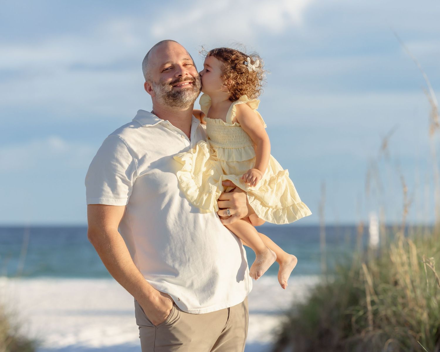 A man holds a young girl in a yellow dress on a sunny beach, both smiling joyfully near ocean waves.