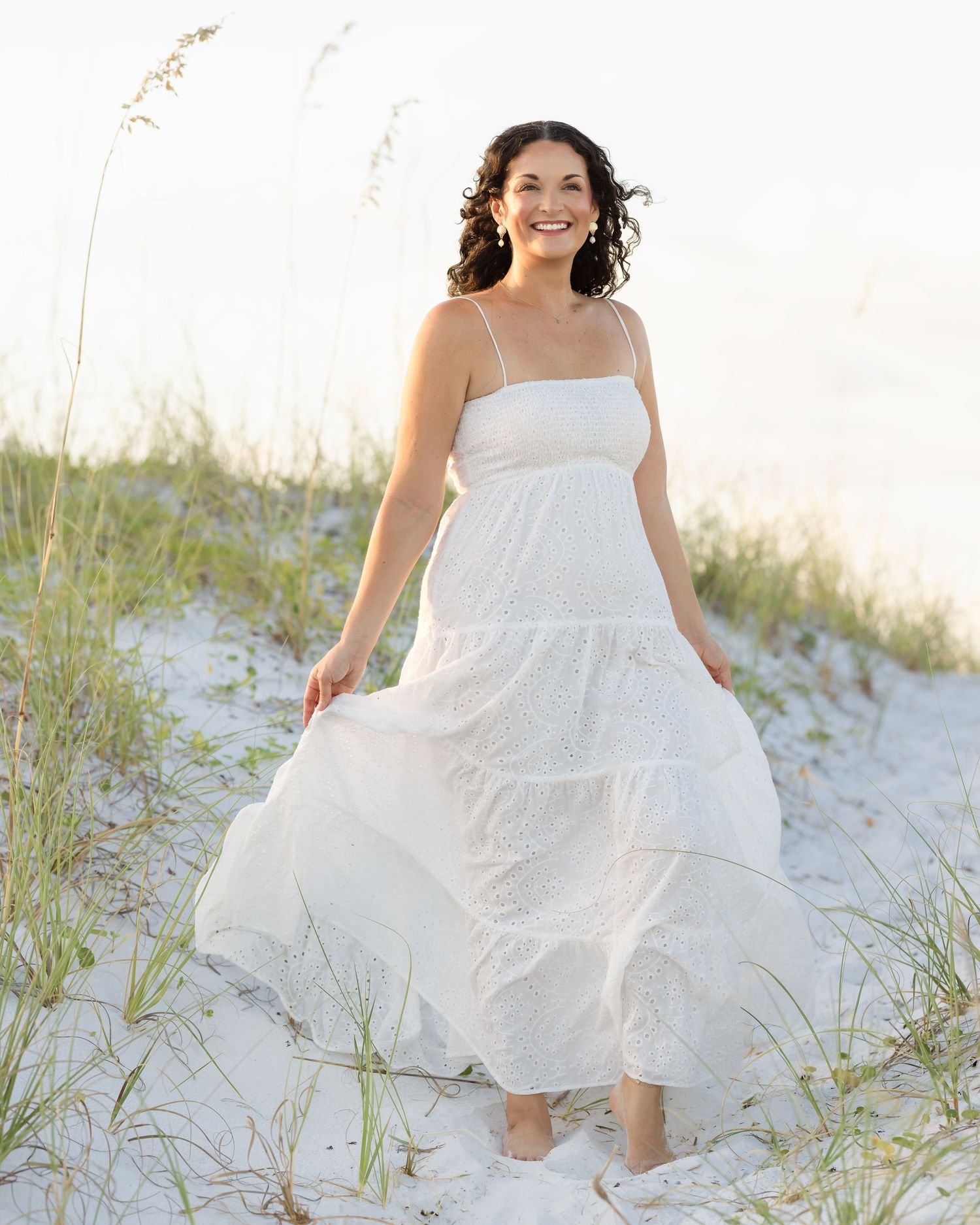 Woman in flowing white sundress smiling on sandy beach dunes.