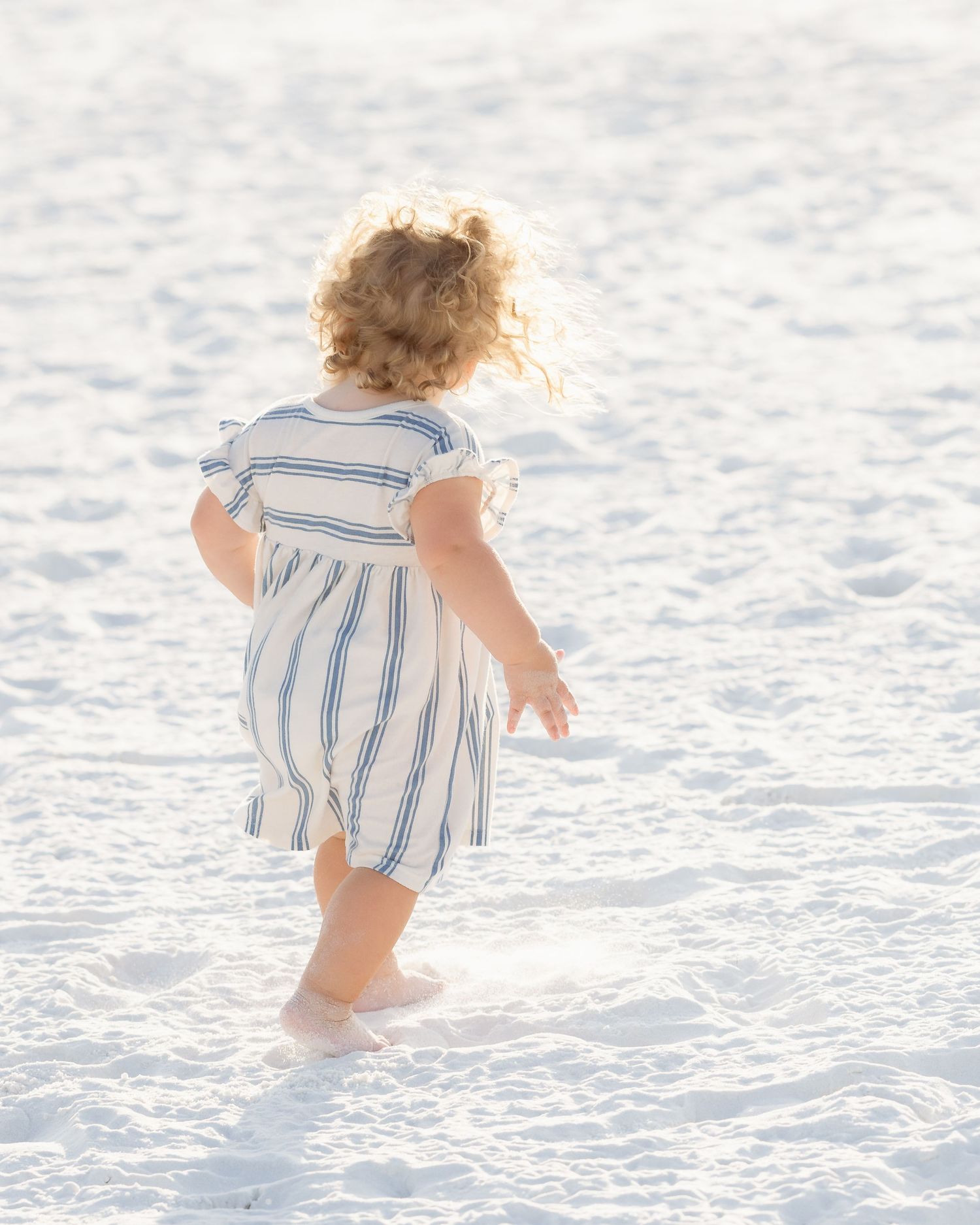 Toddler girl in striped dress walking barefoot on white sand beach.