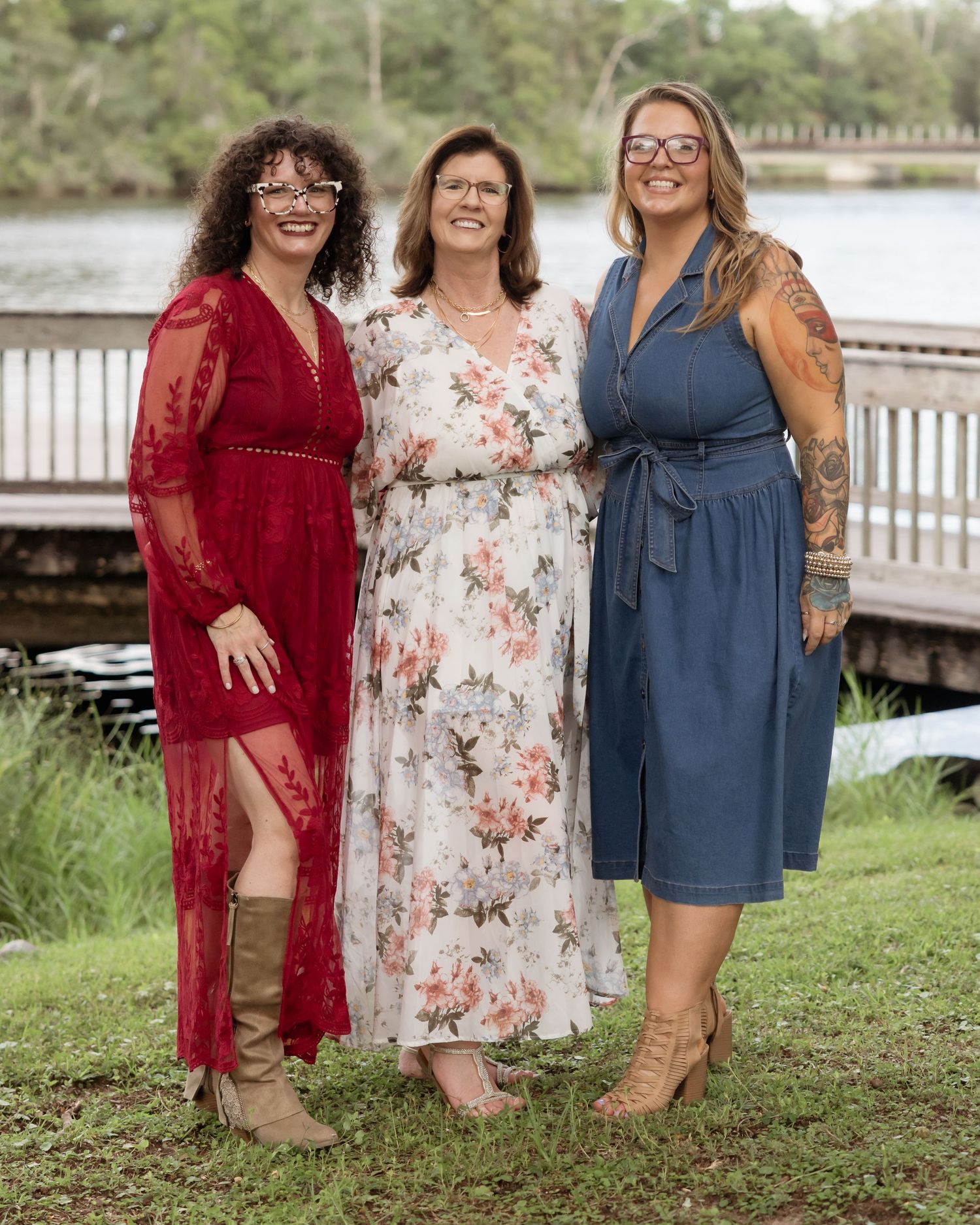 Three women smiling outdoors in elegant dresses near a waterfront dock on a sunny day.