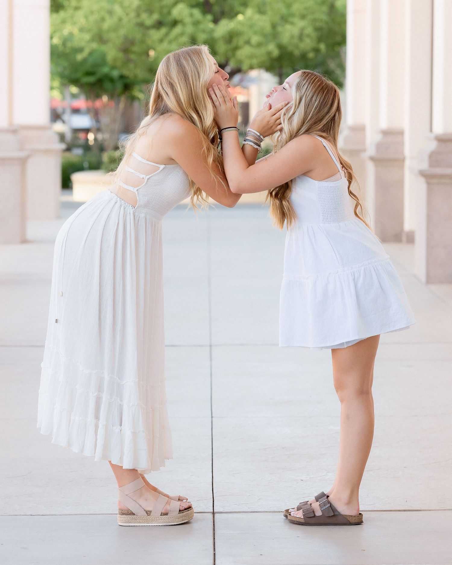Two blonde sisters in white dresses share a sweet kiss on the cheek outdoors.