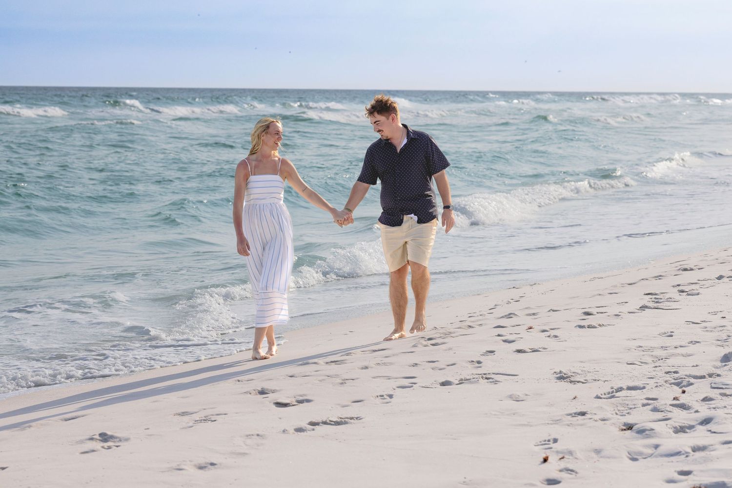 Couple holding hands walking barefoot on a white sandy beach with turquoise ocean waves in the background.