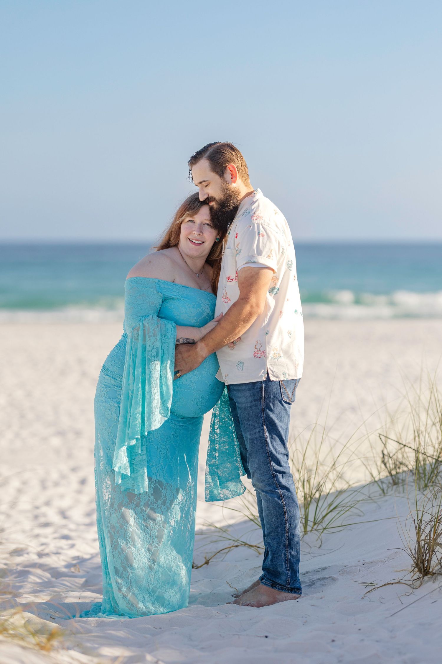 Smiling pregnant woman in blue lace dress cuddled by bearded partner on sunny beach.