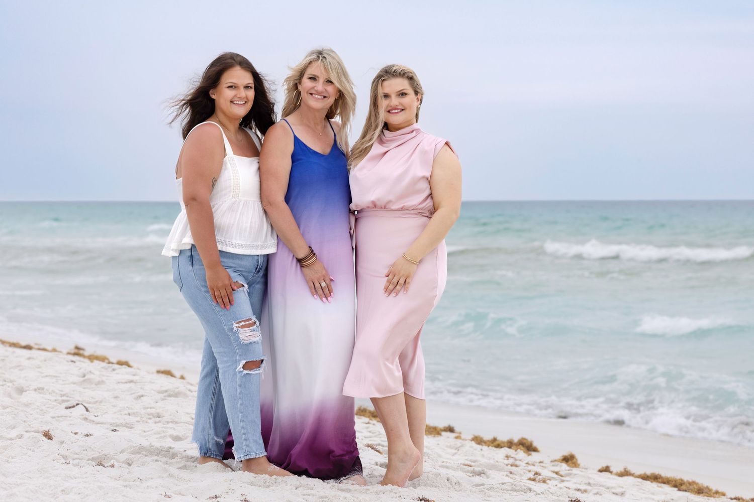 Three women posing together on a sandy beach with ocean waves in the background on a cloudy day.