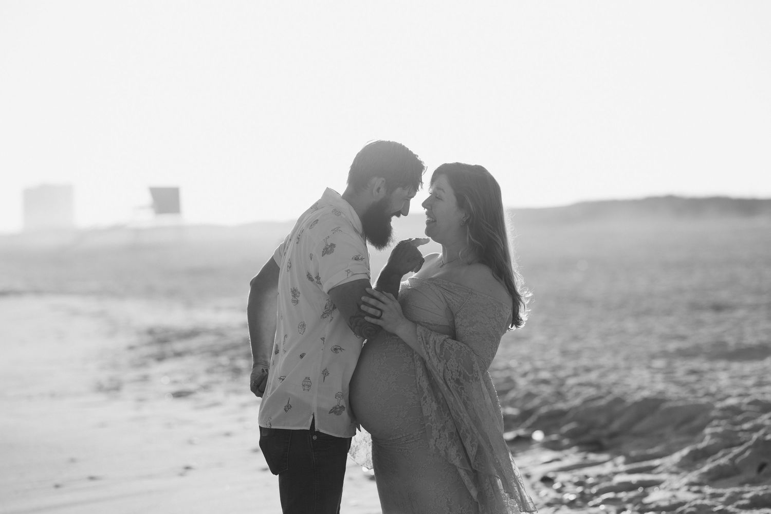 Black and white maternity photo of a couple sharing a tender moment on a beach, woman showing baby bump.