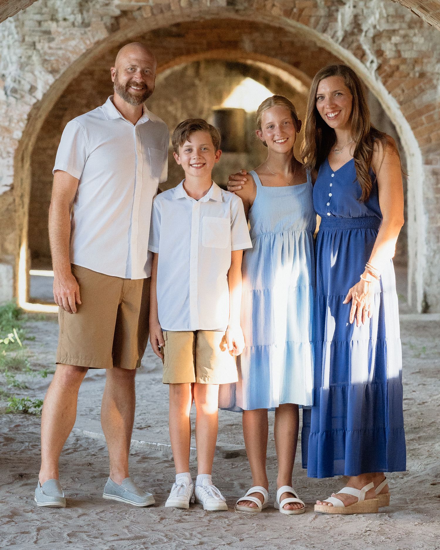 Family of four smiling under stone arches, parents and two children dressed in blue and white summer outfits.