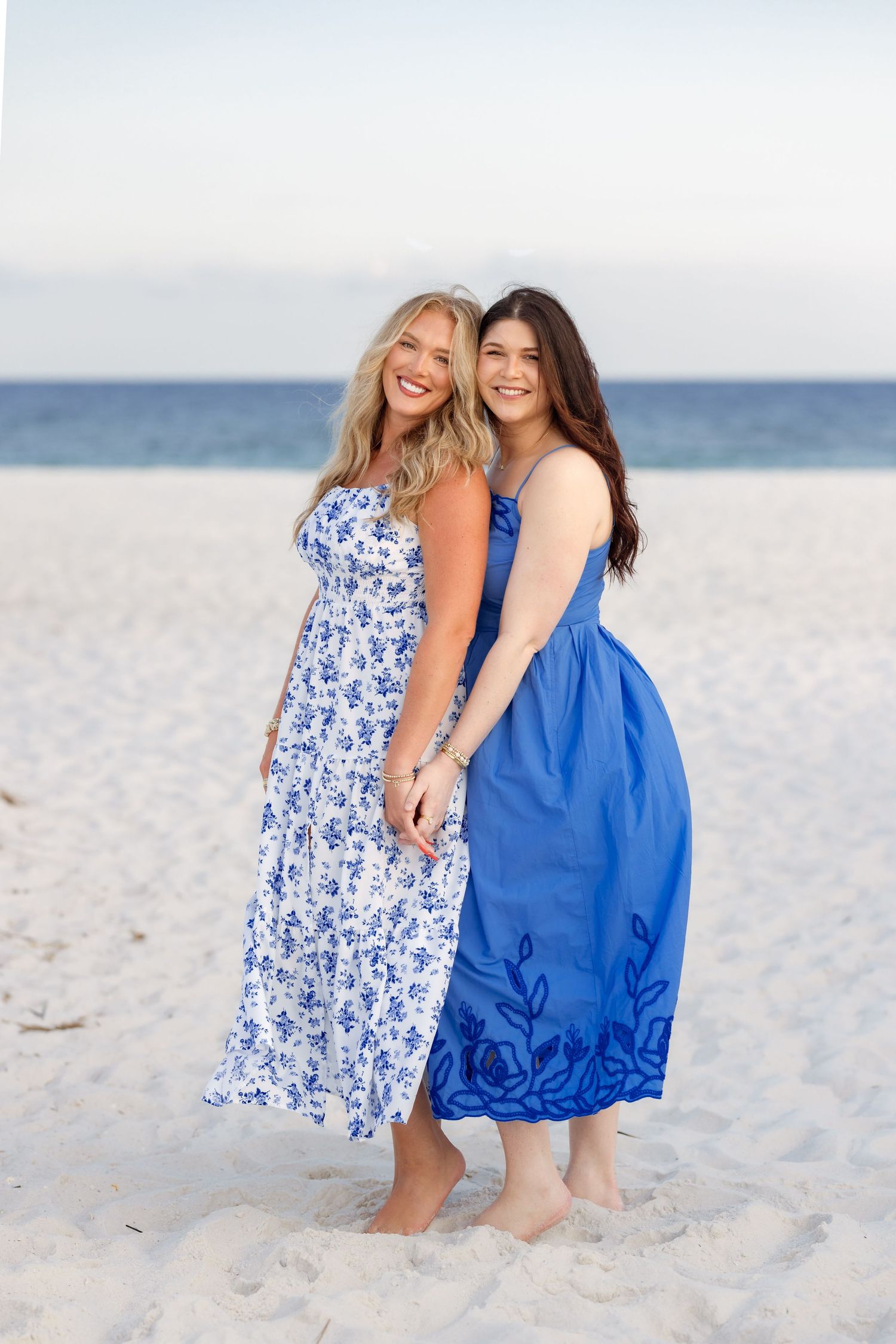 Two young women in blue dresses smiling on a sandy beach with ocean in background.