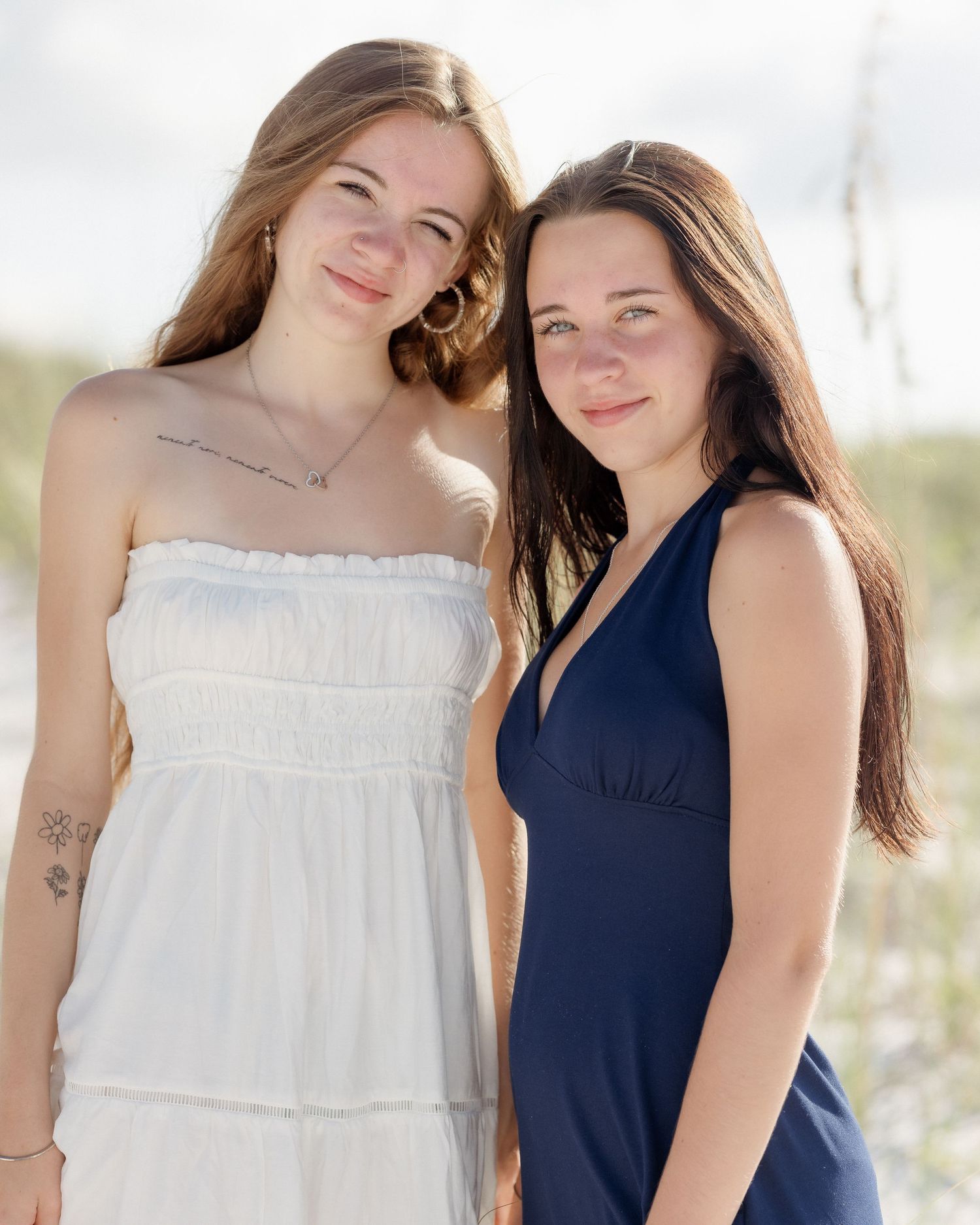 Two young women posing together on a beach, one in a white dress and one in a navy blue dress.