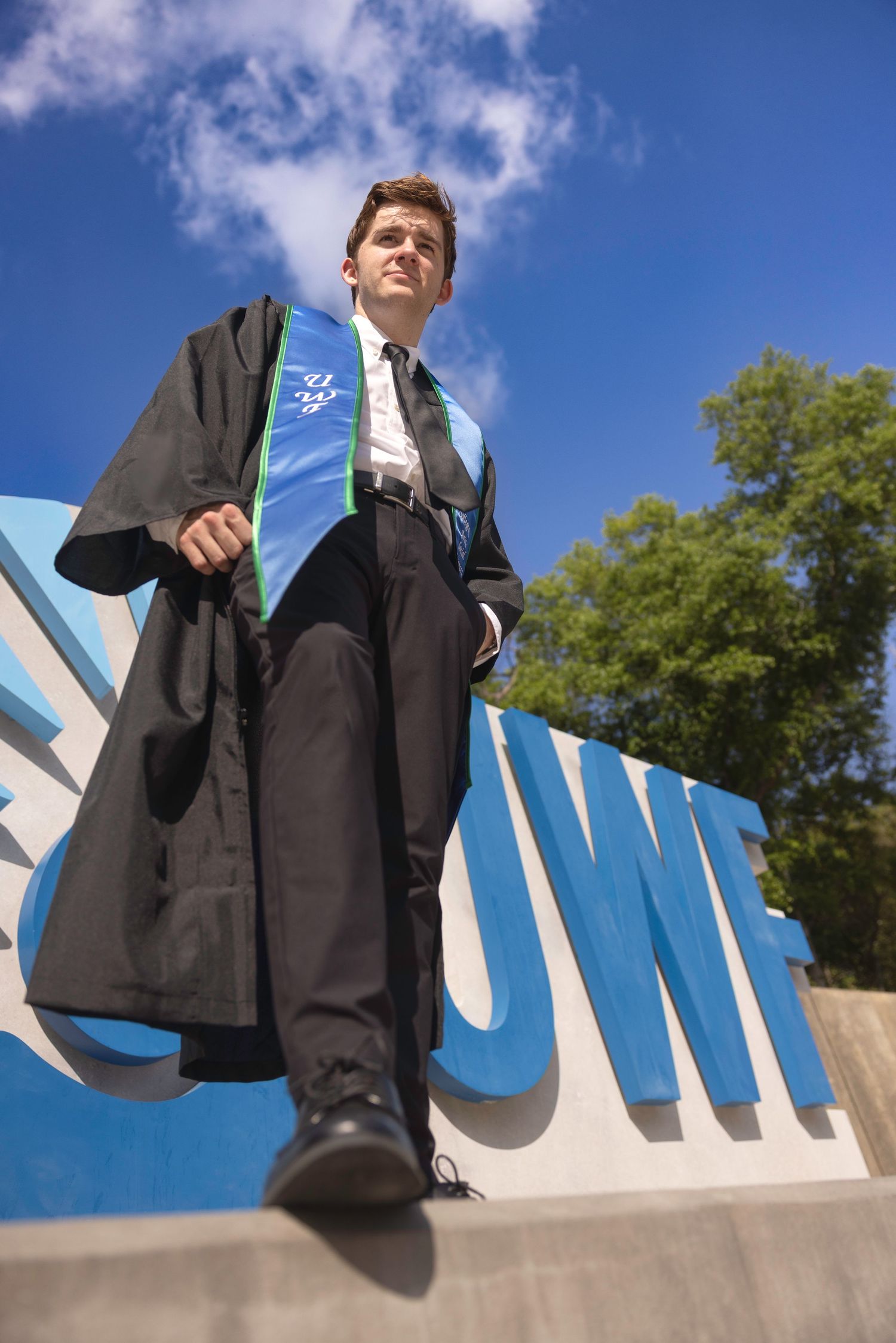 Graduate in black gown and green sash poses confidently against large blue university sign under bright sky.