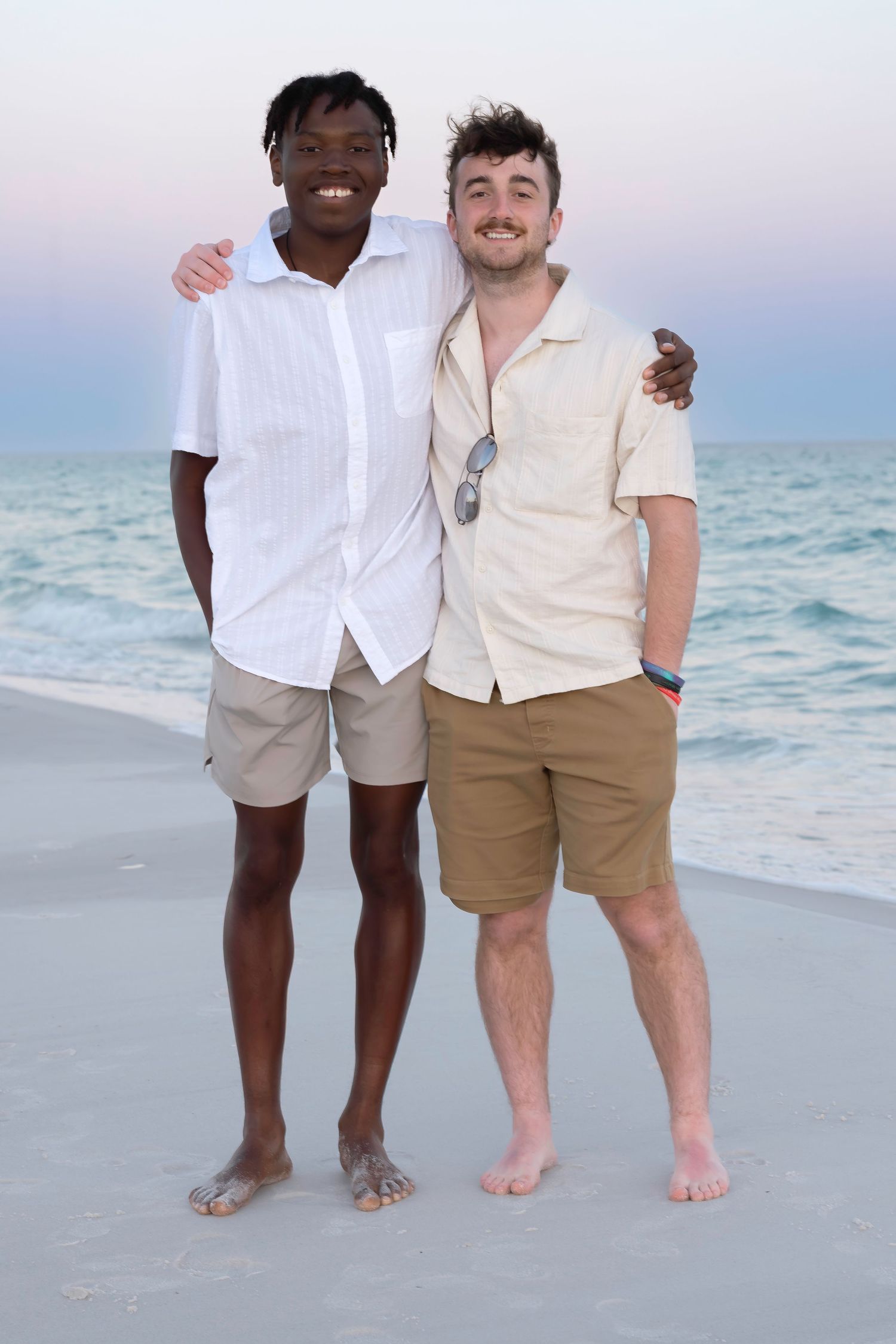 Two men smiling and posing together on a beach at sunset, wearing casual shirts and khaki shorts.