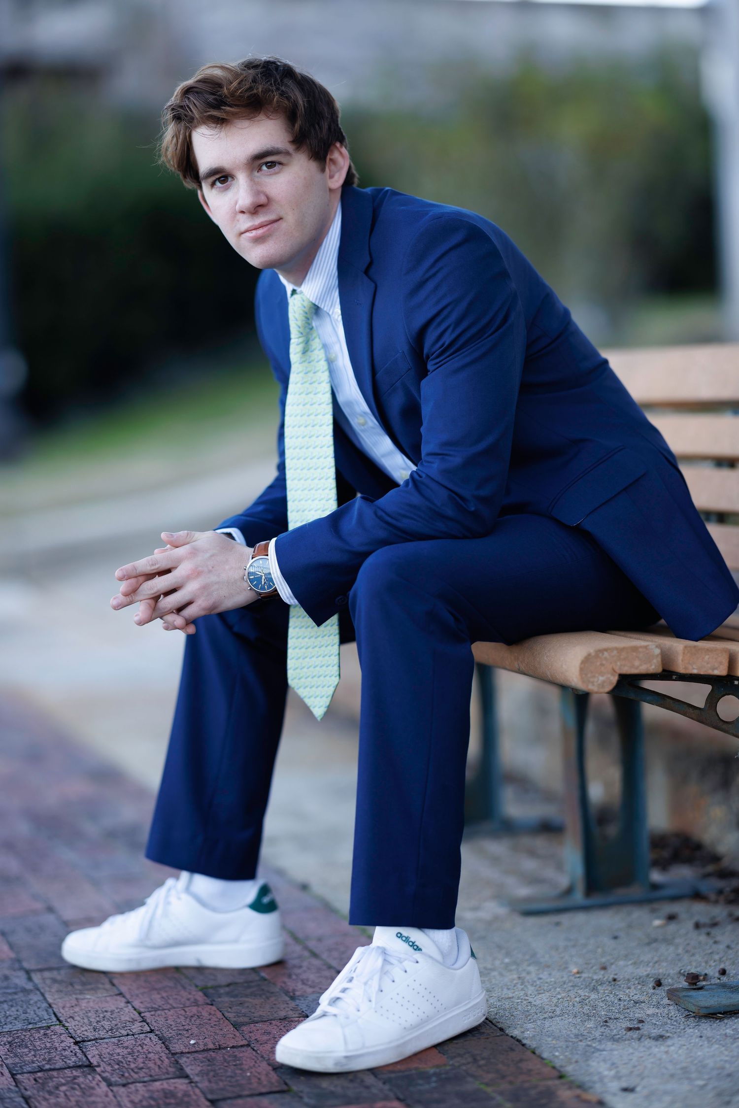 Young man in navy blue suit and white sneakers sitting on a bench outdoors.