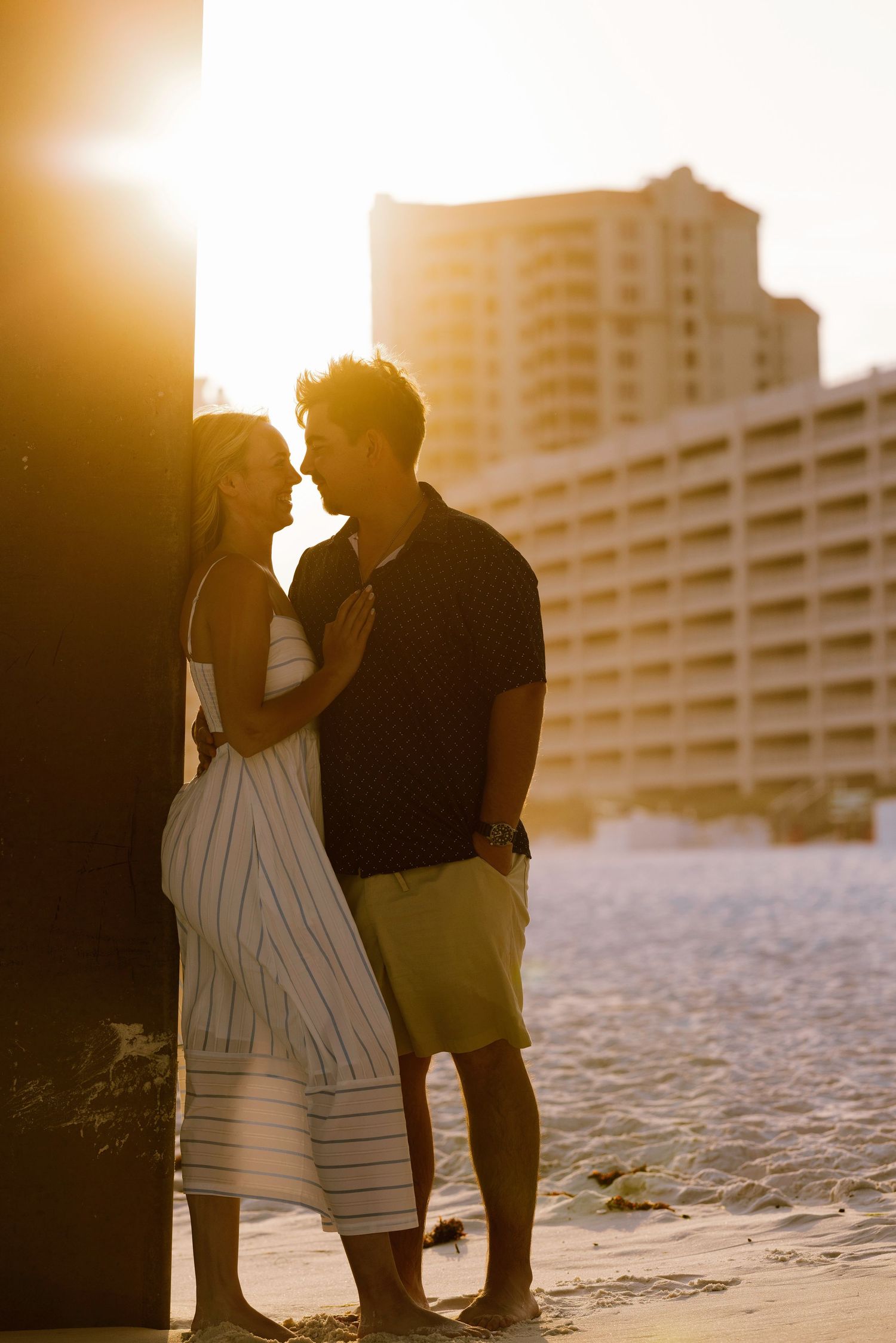 Couple sharing a romantic moment at sunset on a sandy beach with a resort building in the background.