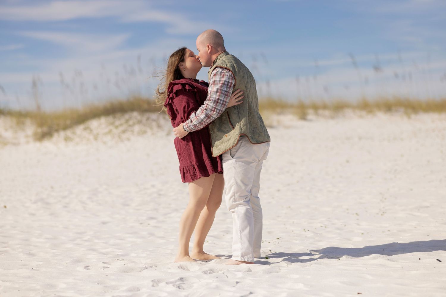 Couple embracing on a pristine white sandy beach with soft blue sky in the background.