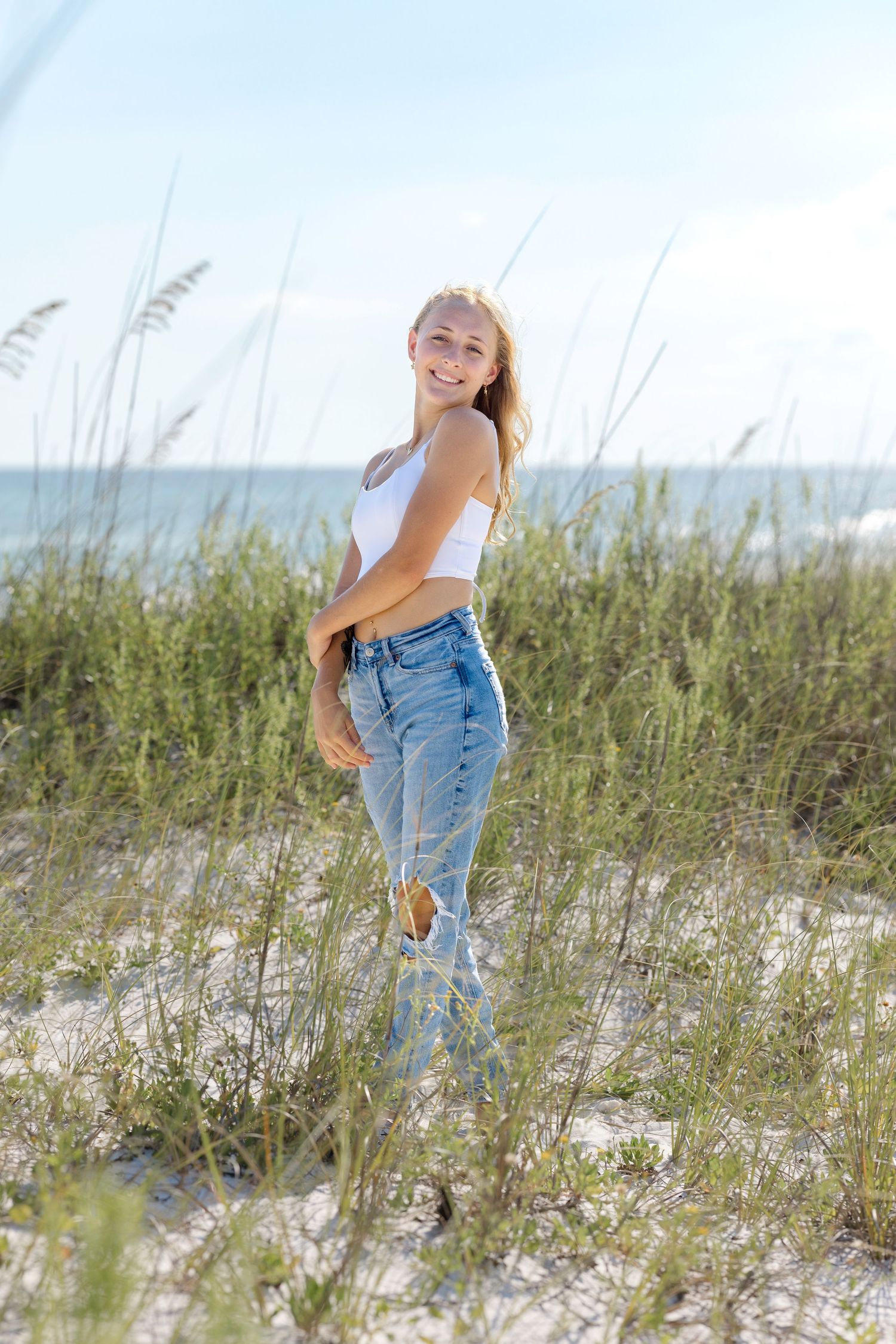 A person in casual denim and white attire poses playfully among beach grass and sand dunes on a sunny day.
