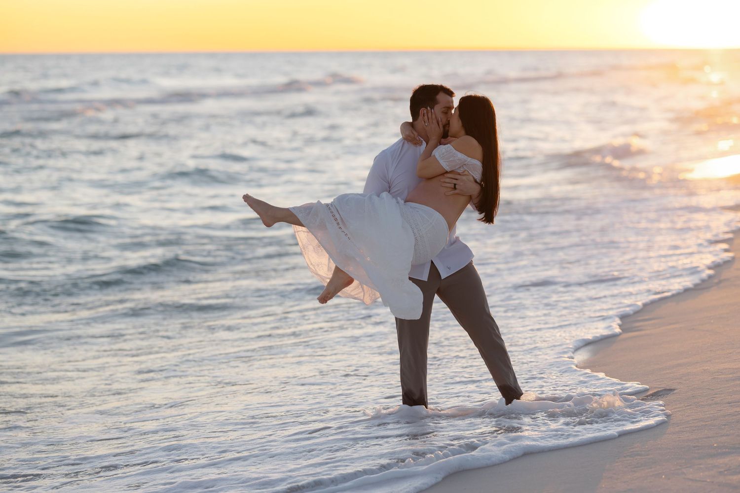 Couple in white clothing embracing and dancing on a beach at sunset with ocean waves in the background.