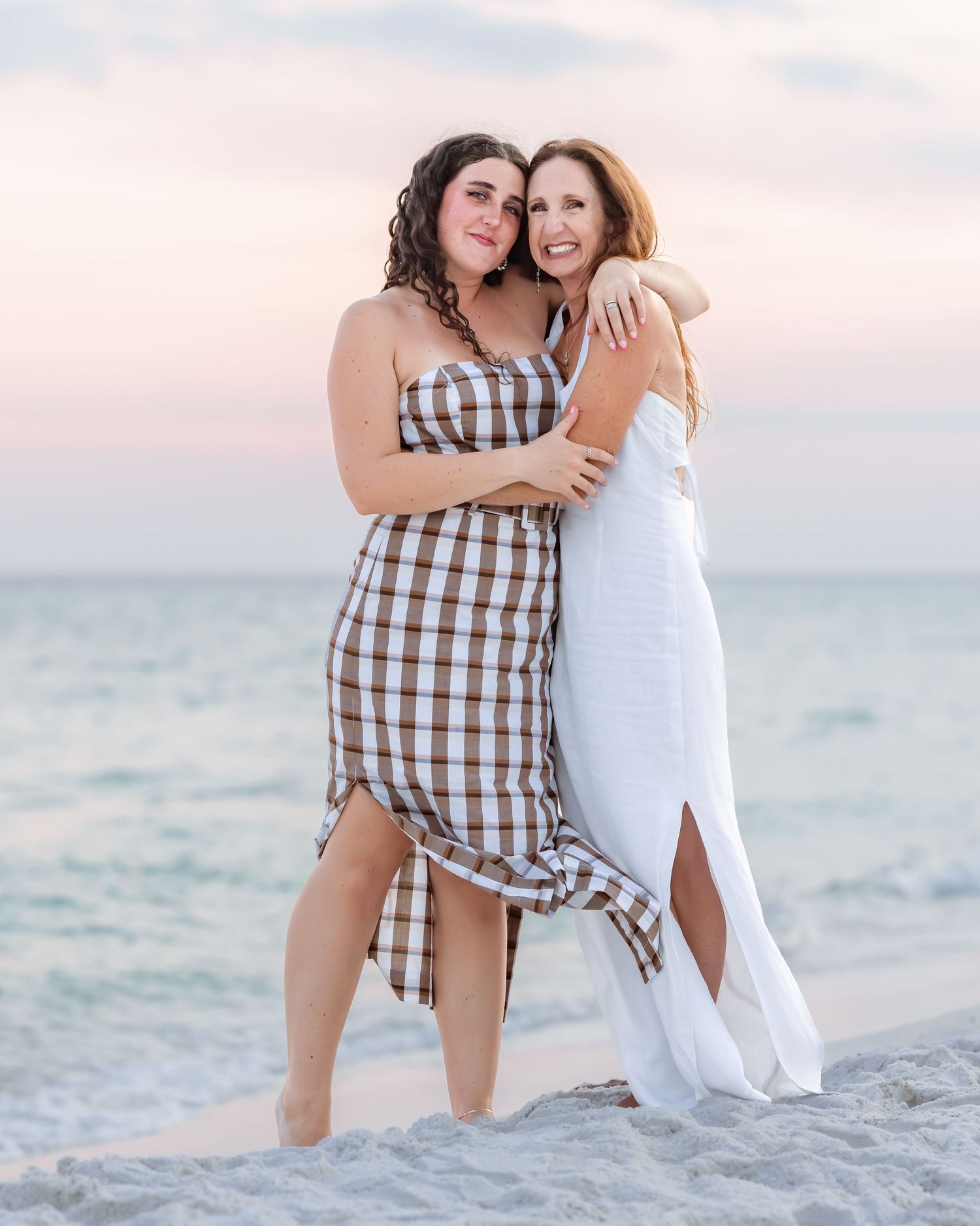 Mother and teenage daughter embrace warmly on sandy beach at sunset in coordinating stylish outfits.