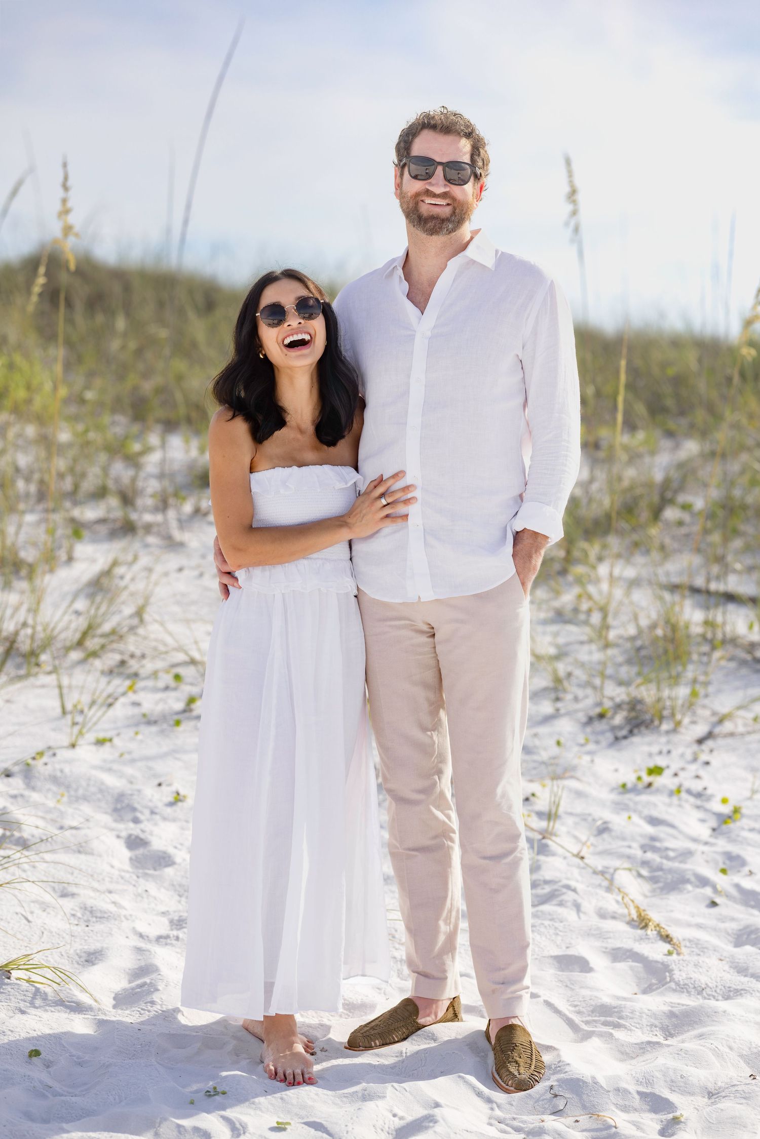 Happy couple in white outfits and sunglasses posing on sandy beach dunes with sea oats in background.