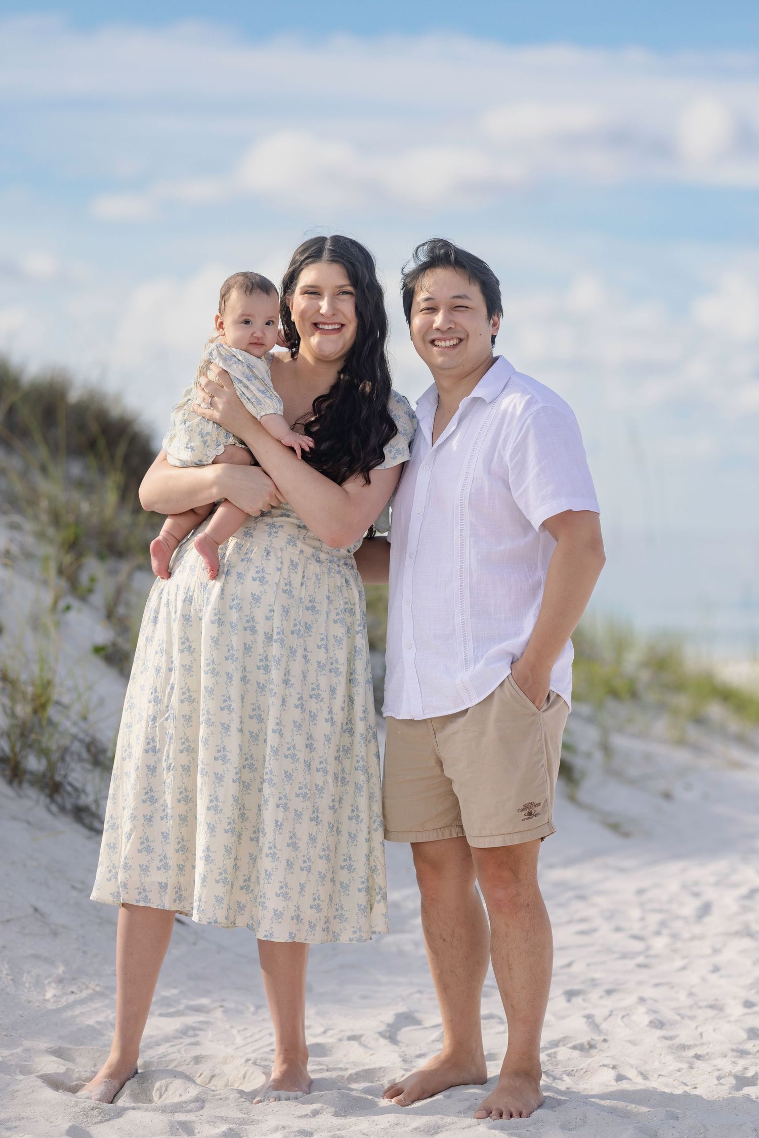 A young family with a baby poses barefoot on a white sand beach with dunes and blue sky behind them.