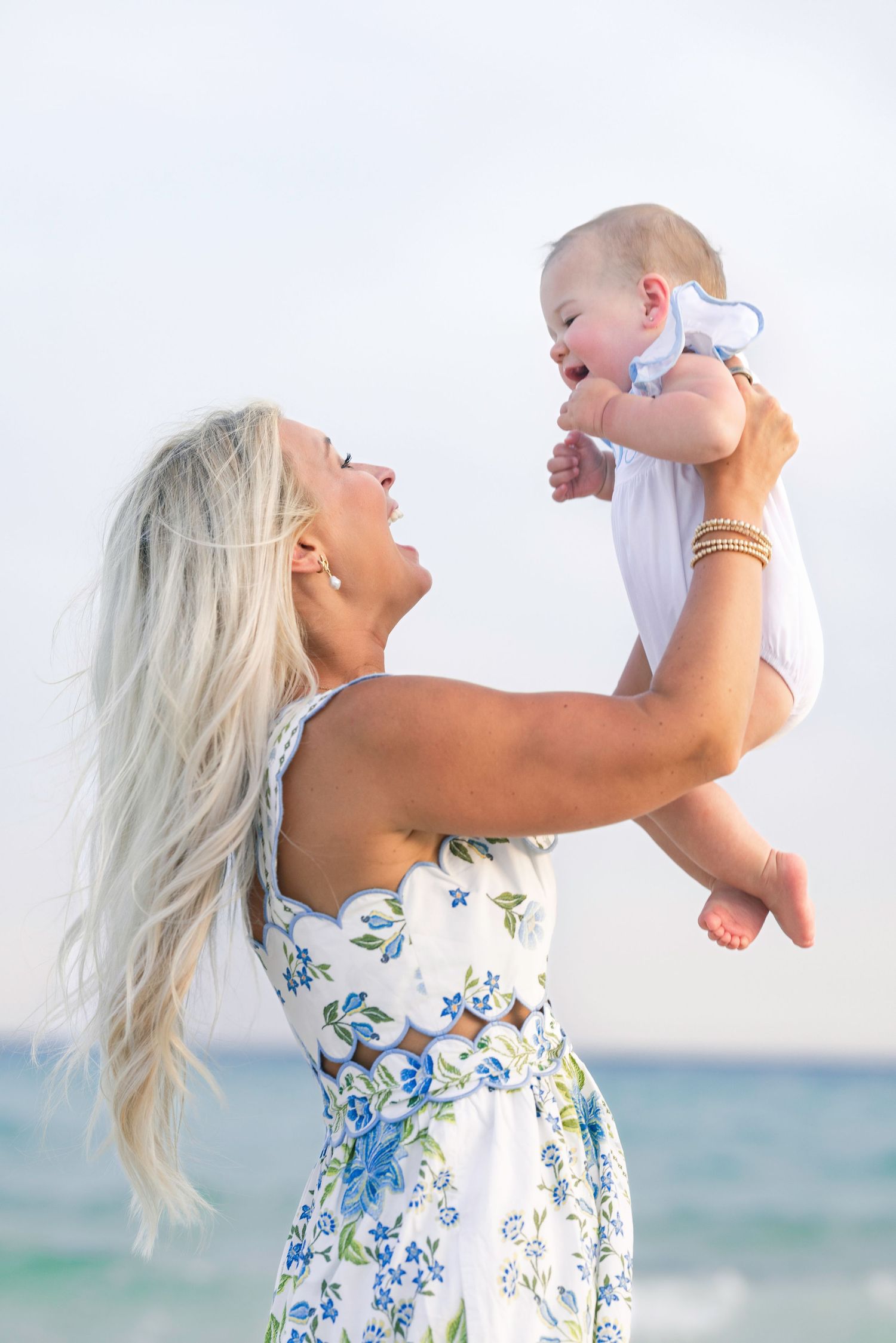 Blonde woman in floral dress joyfully lifts a smiling baby at the beach.