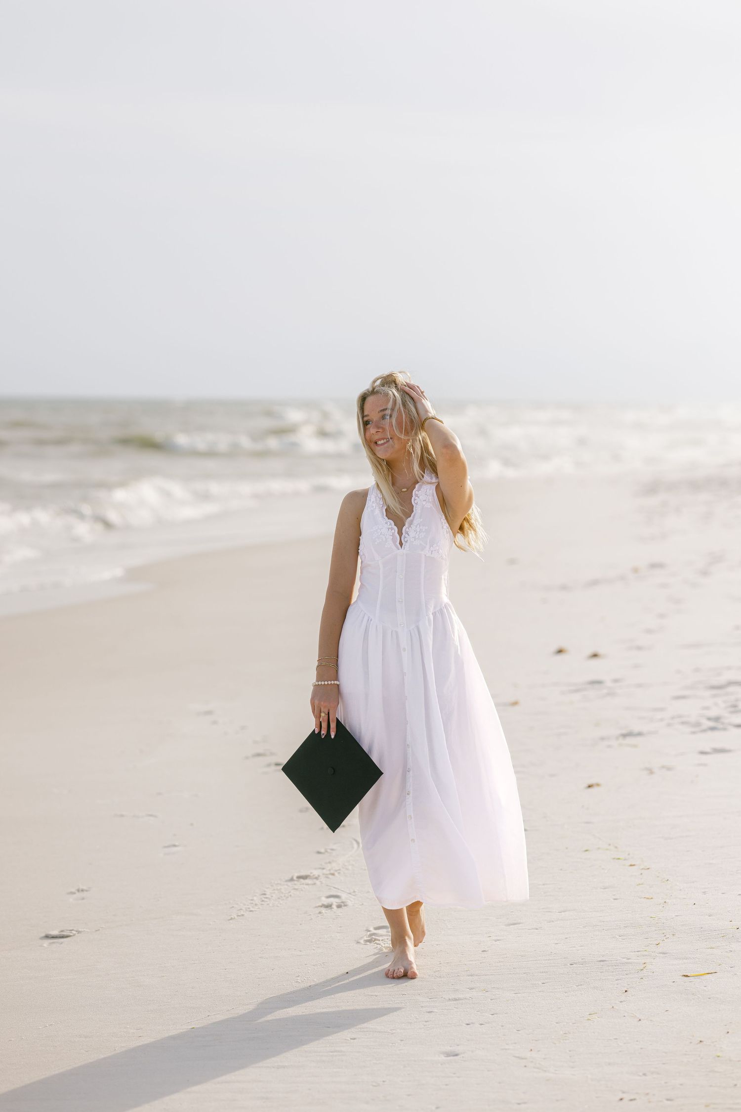 Blonde woman in white maxi dress walking barefoot on beach holding black graduation cap.