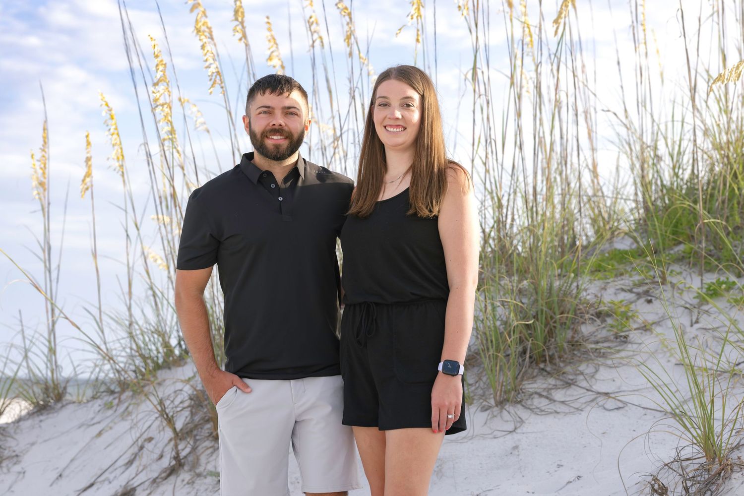 A couple in matching black outfits poses together on a sandy beach with sea oats and dunes in the background.