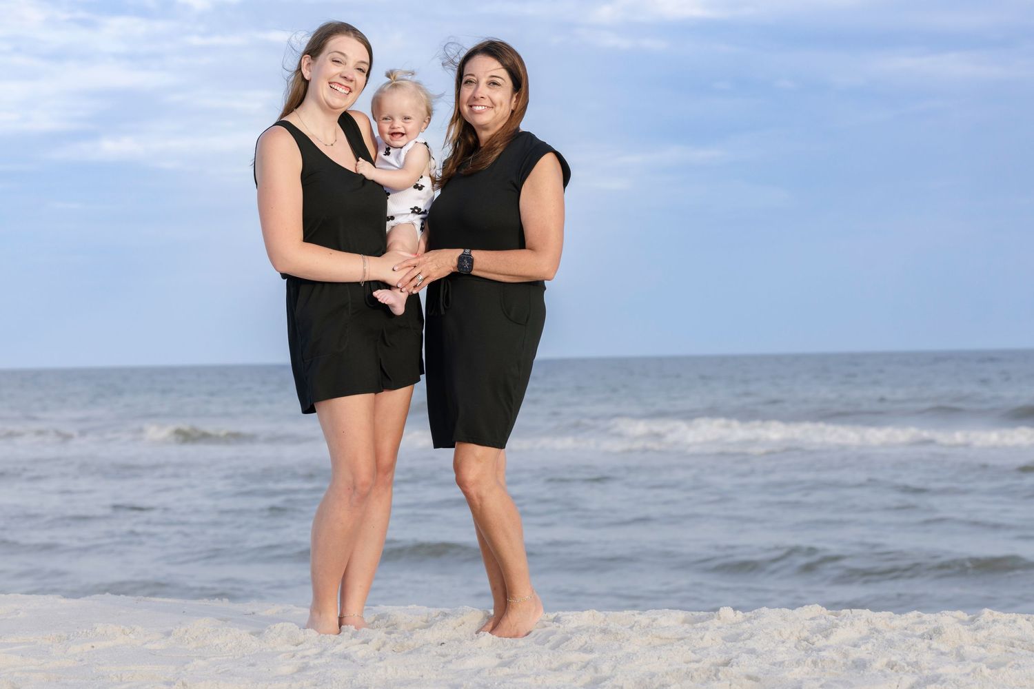 Two women in black dresses smile on a sunny beach, one holding a happy baby in white.