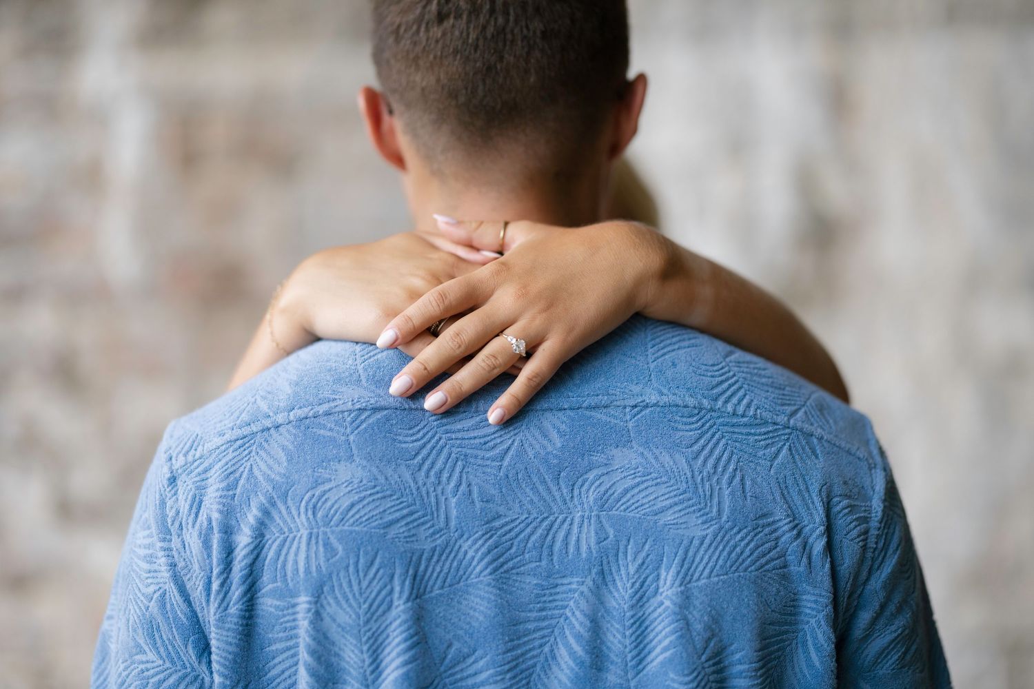 A woman's hand with an engagement ring resting on a man's shoulder in a blue patterned shirt.