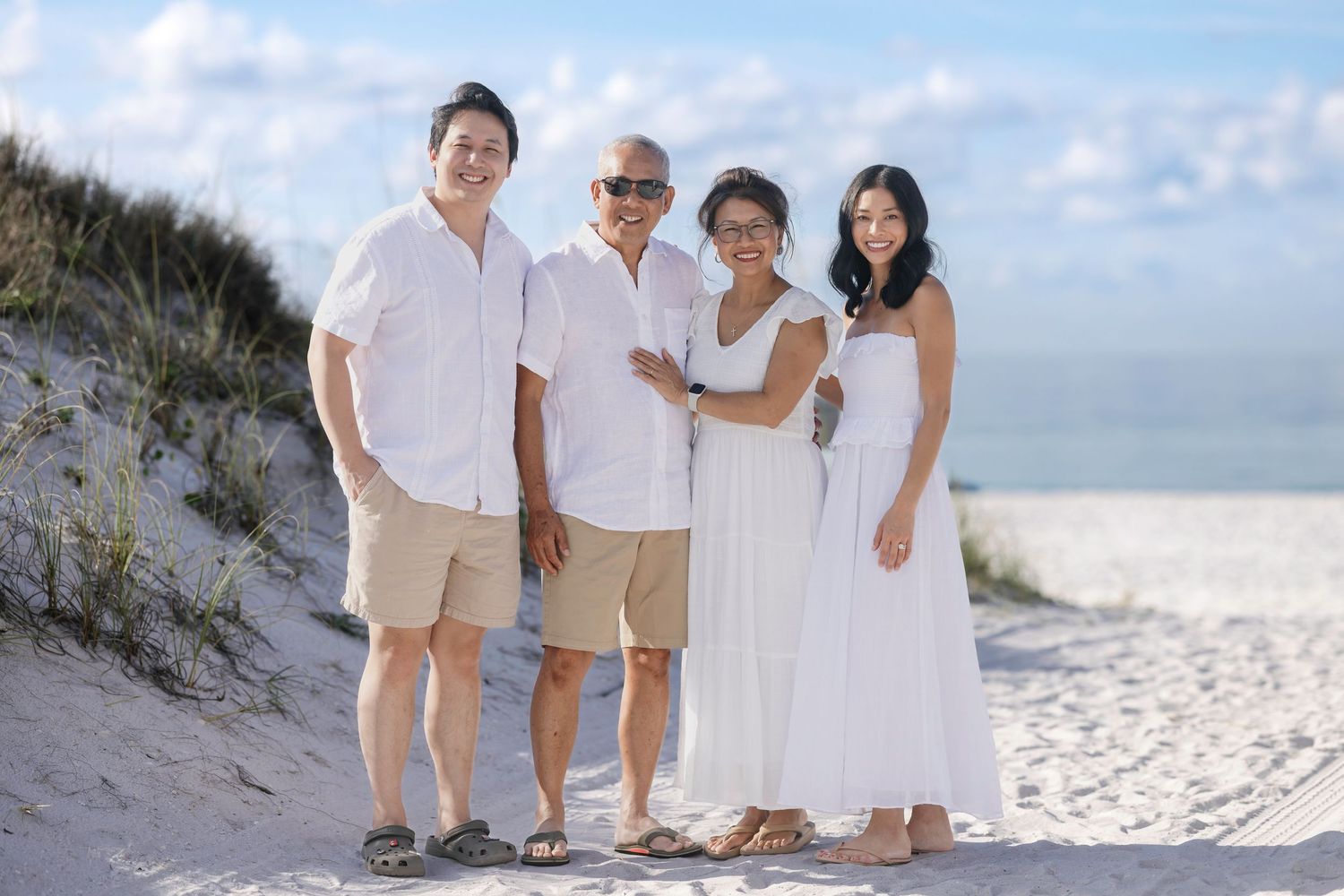 Family of four posing together on a sandy beach, dressed in white and khaki outfits, with dunes and blue sky behind them.