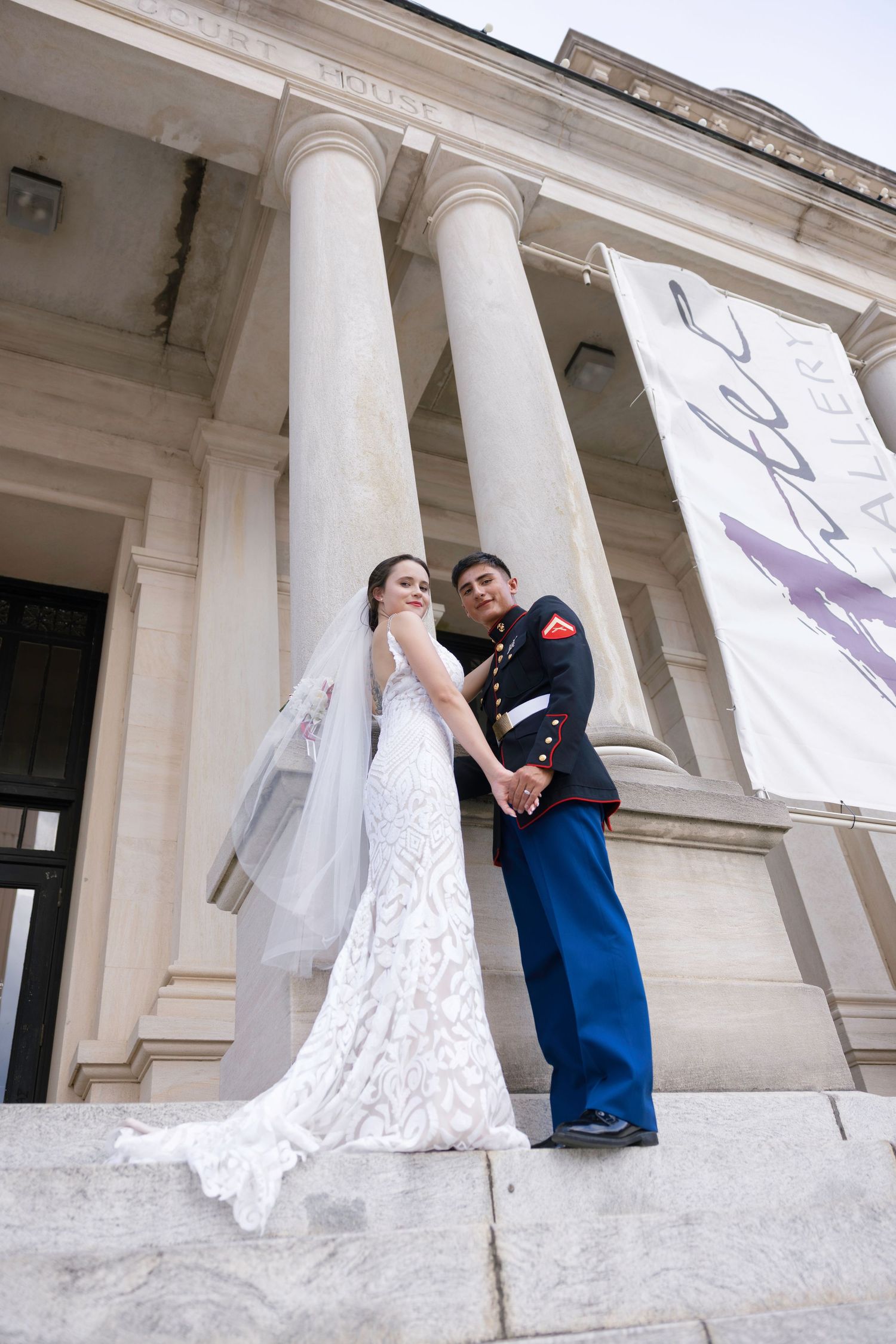 A bride in white gown and groom in Marine dress blues pose on steps of a neoclassical building.