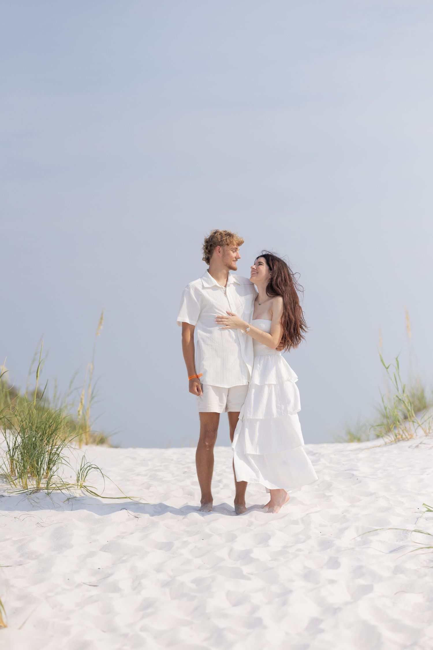 Couple in white outfits walking barefoot on white sand beach dunes, gazing at each other romantically.