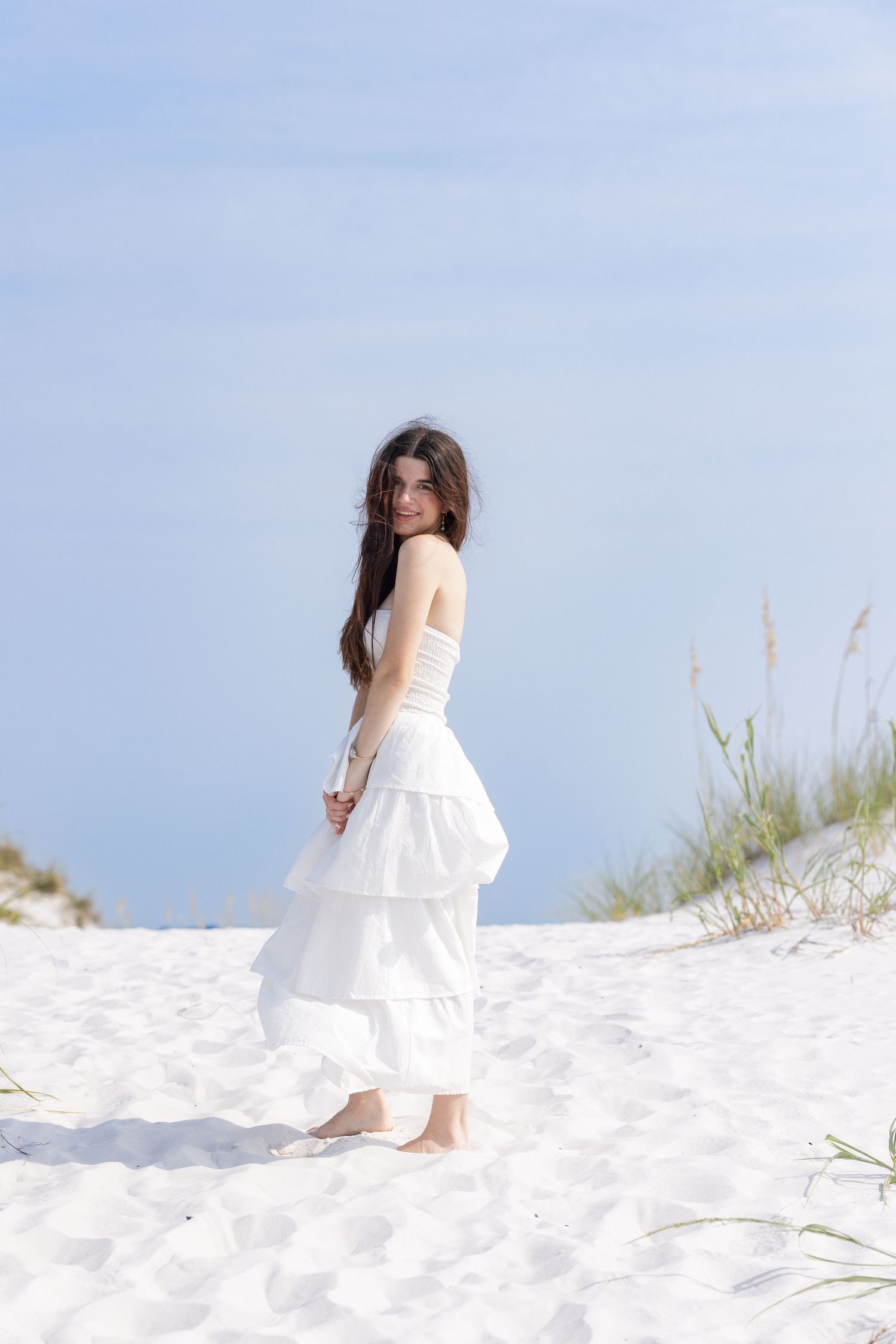 Young woman in white strapless tiered dress standing barefoot on white sand beach dunes under blue sky.