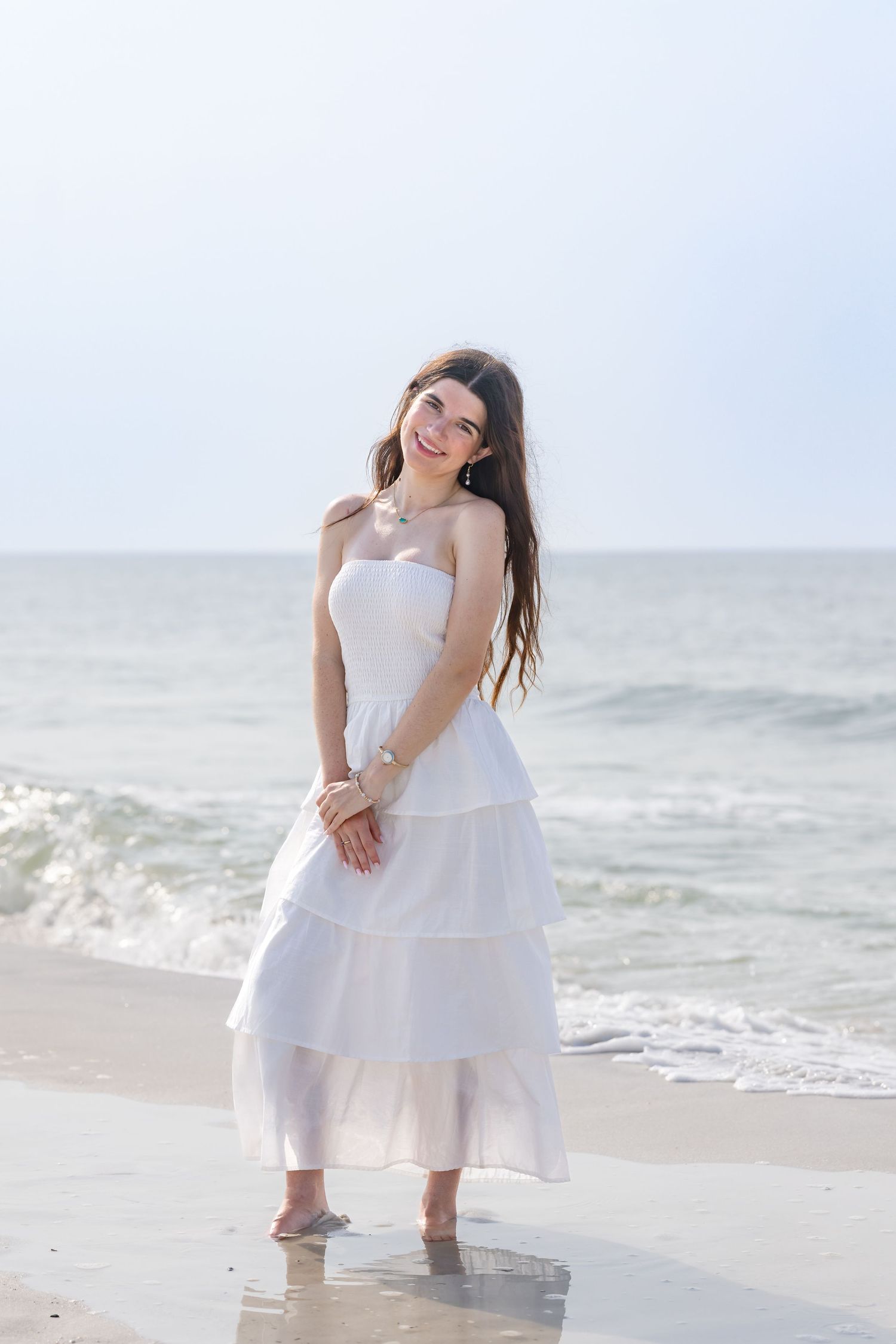 Smiling young woman in white strapless tiered dress standing at ocean shoreline with waves behind her.