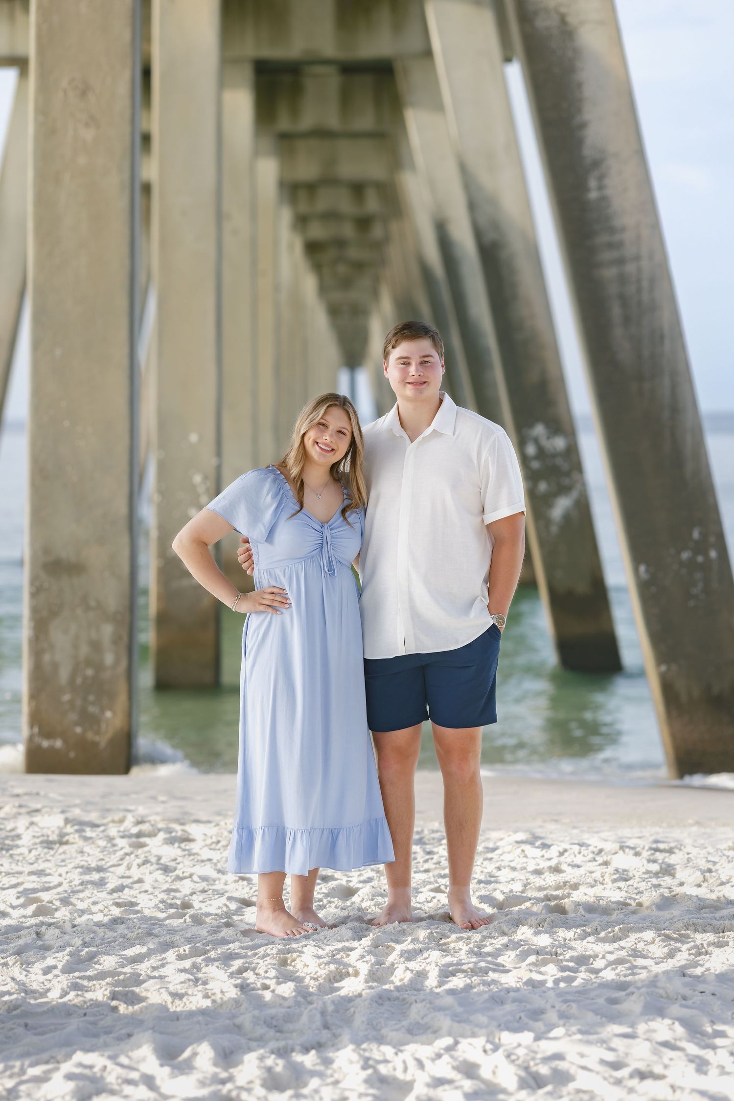 A smiling couple poses barefoot on white sand beach under a pier, wearing light blue dress and white shirt with navy shorts.