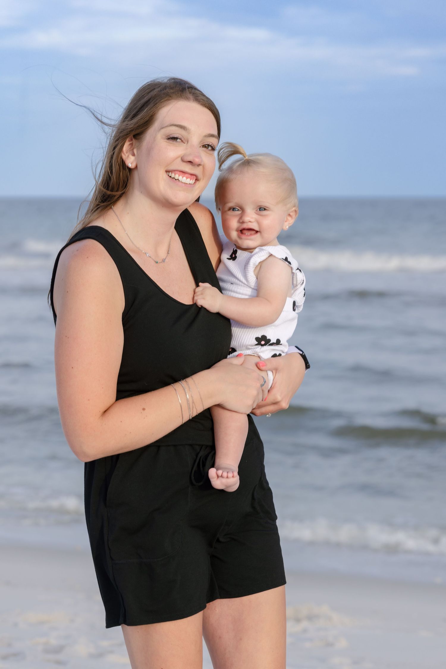 Smiling young woman in black romper holding a baby in white outfit on a sunny beach with ocean waves behind them.