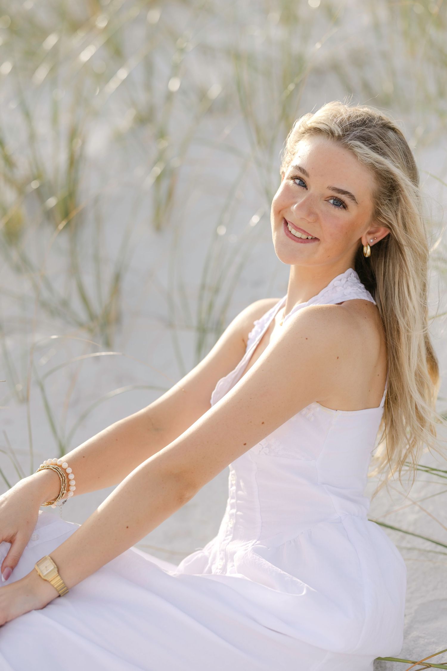 Young blonde woman in white dress sitting on sandy beach dunes, smiling outdoors in natural sunlight.