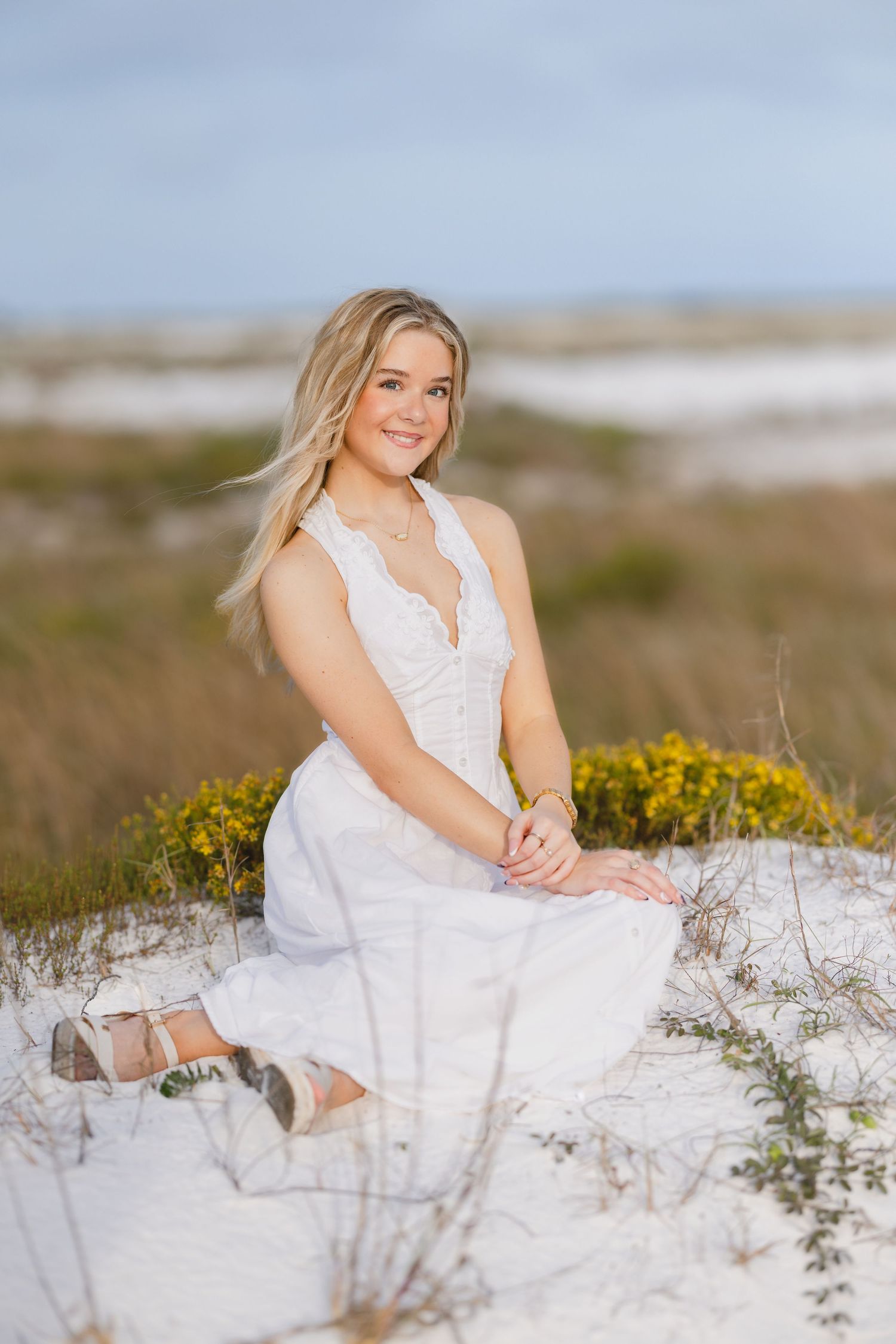Young blonde woman in white lace dress sitting on sandy beach dunes with yellow wildflowers, smiling outdoors.