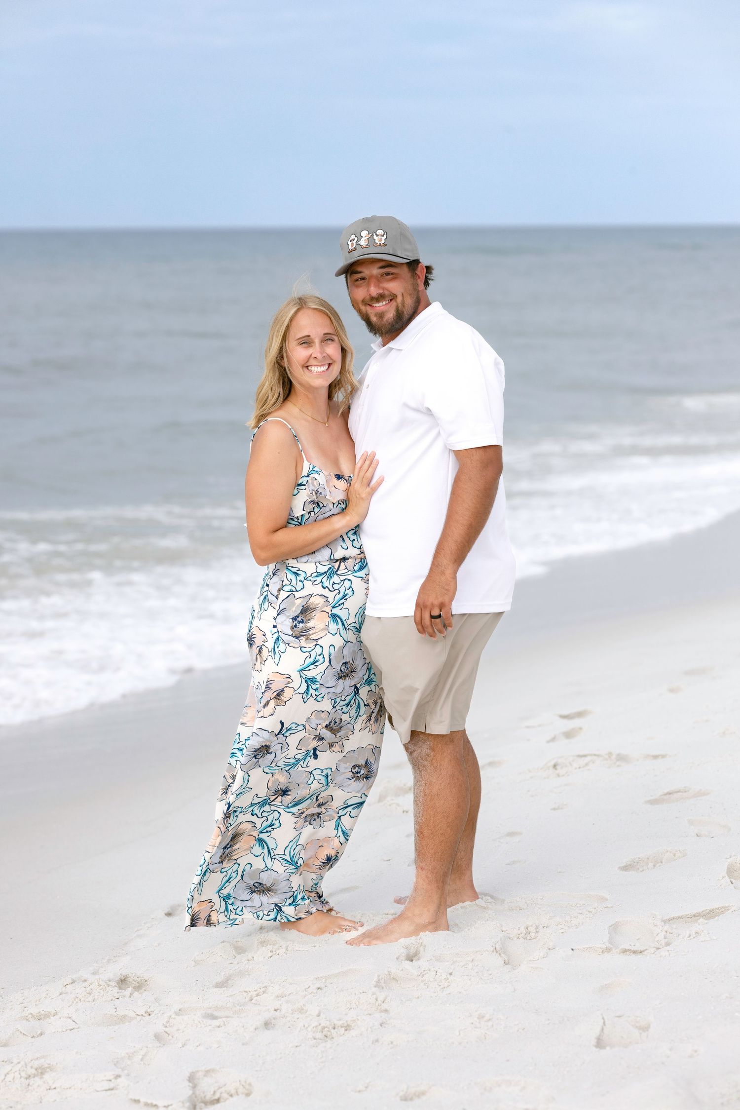 A smiling couple poses on a white sandy beach, woman in floral maxi dress, man in white shirt and cap, ocean backdrop.