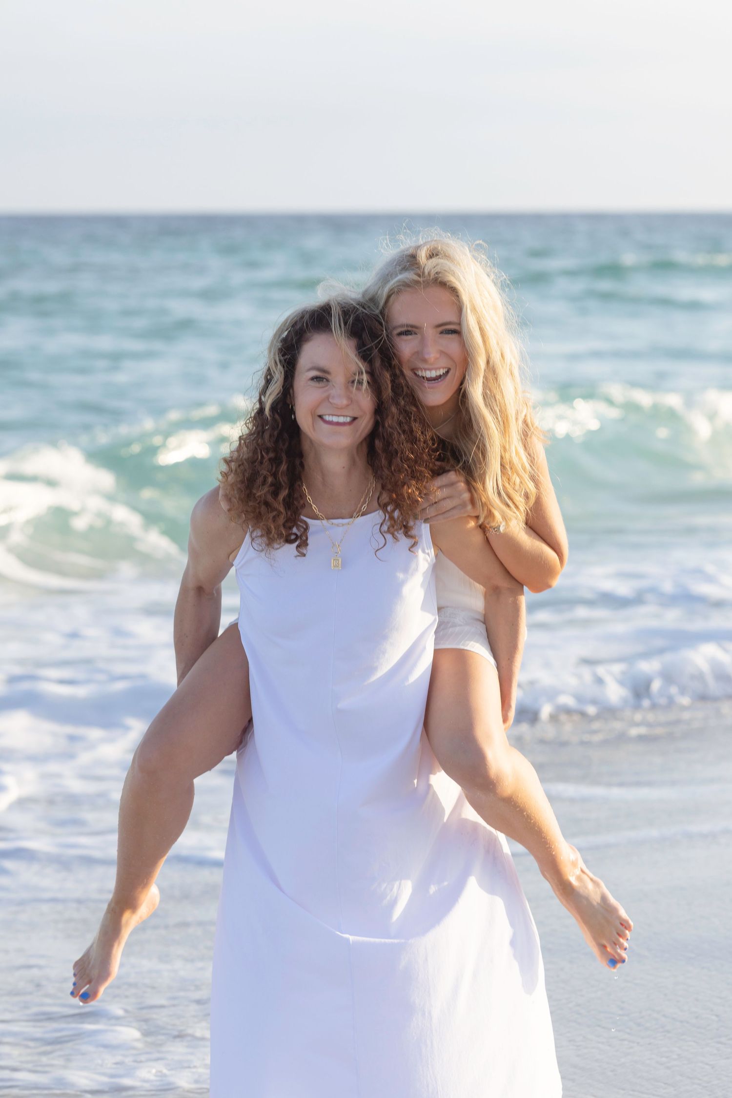 Two women laughing on a beach, one giving the other a piggyback ride in the ocean waves.