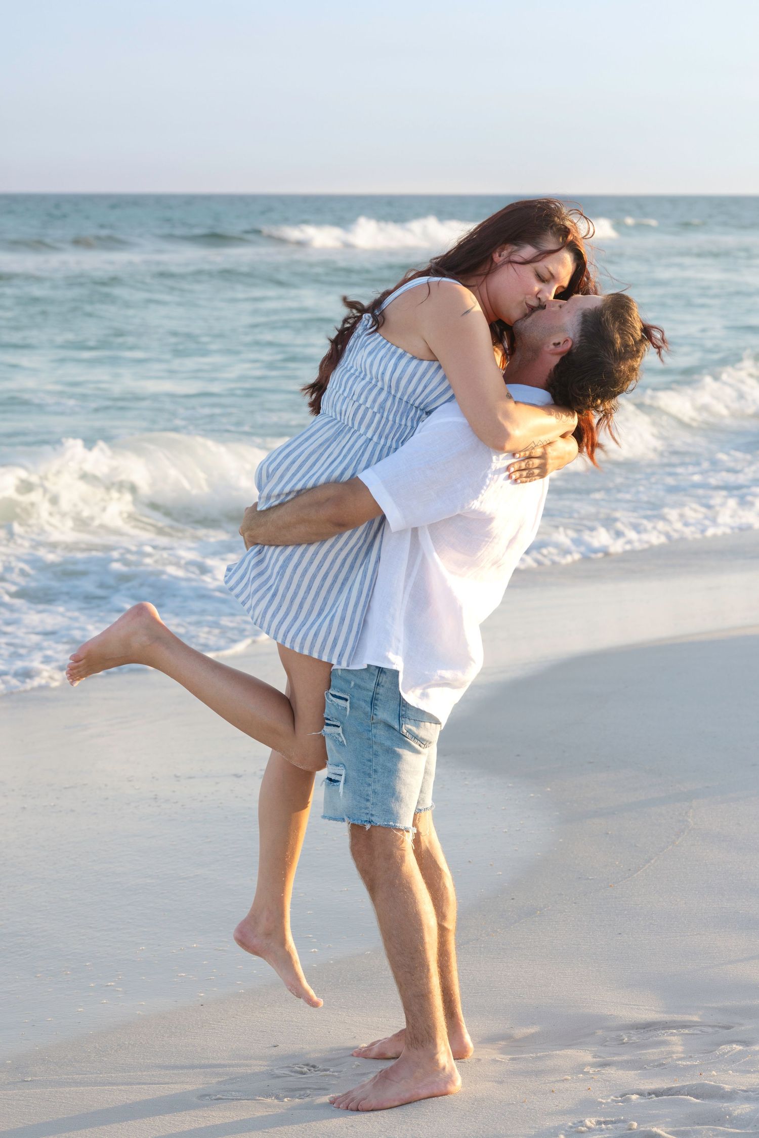 Romantic couple kissing on the beach as man lifts woman near ocean waves at sunset.