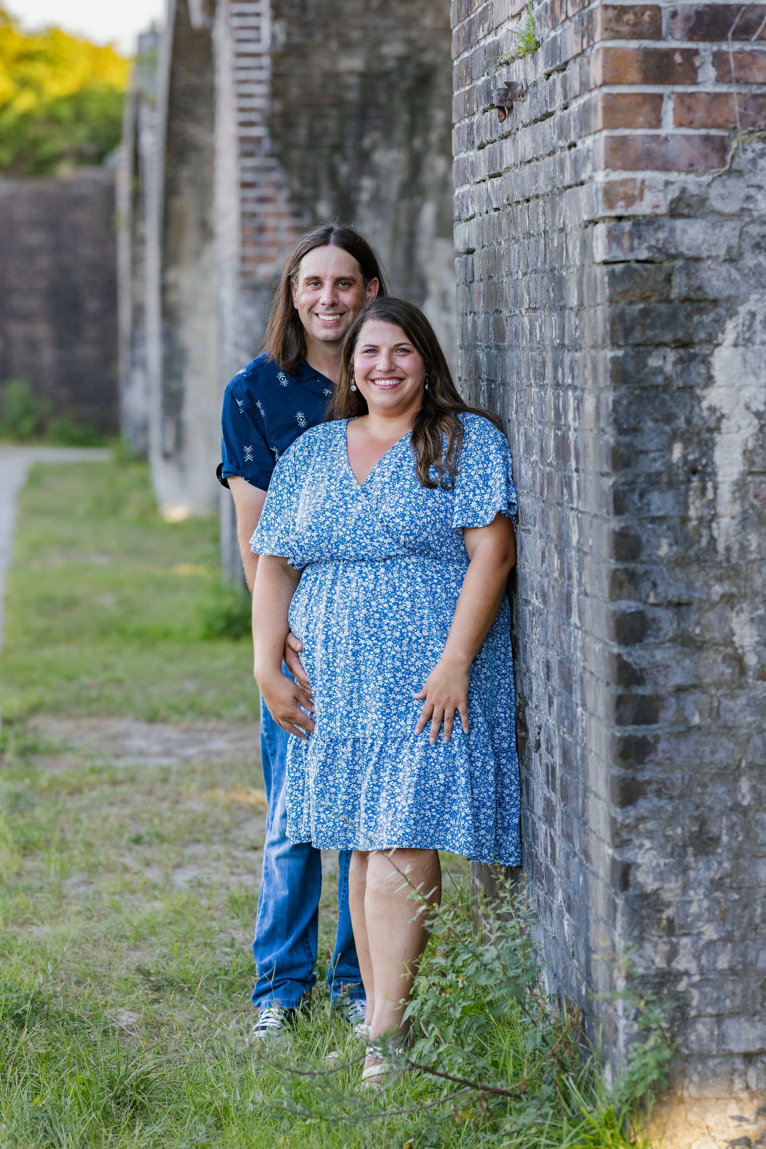 Happy couple posing beside a rustic brick wall outdoors, woman in a blue floral dress, man in a blue shirt.