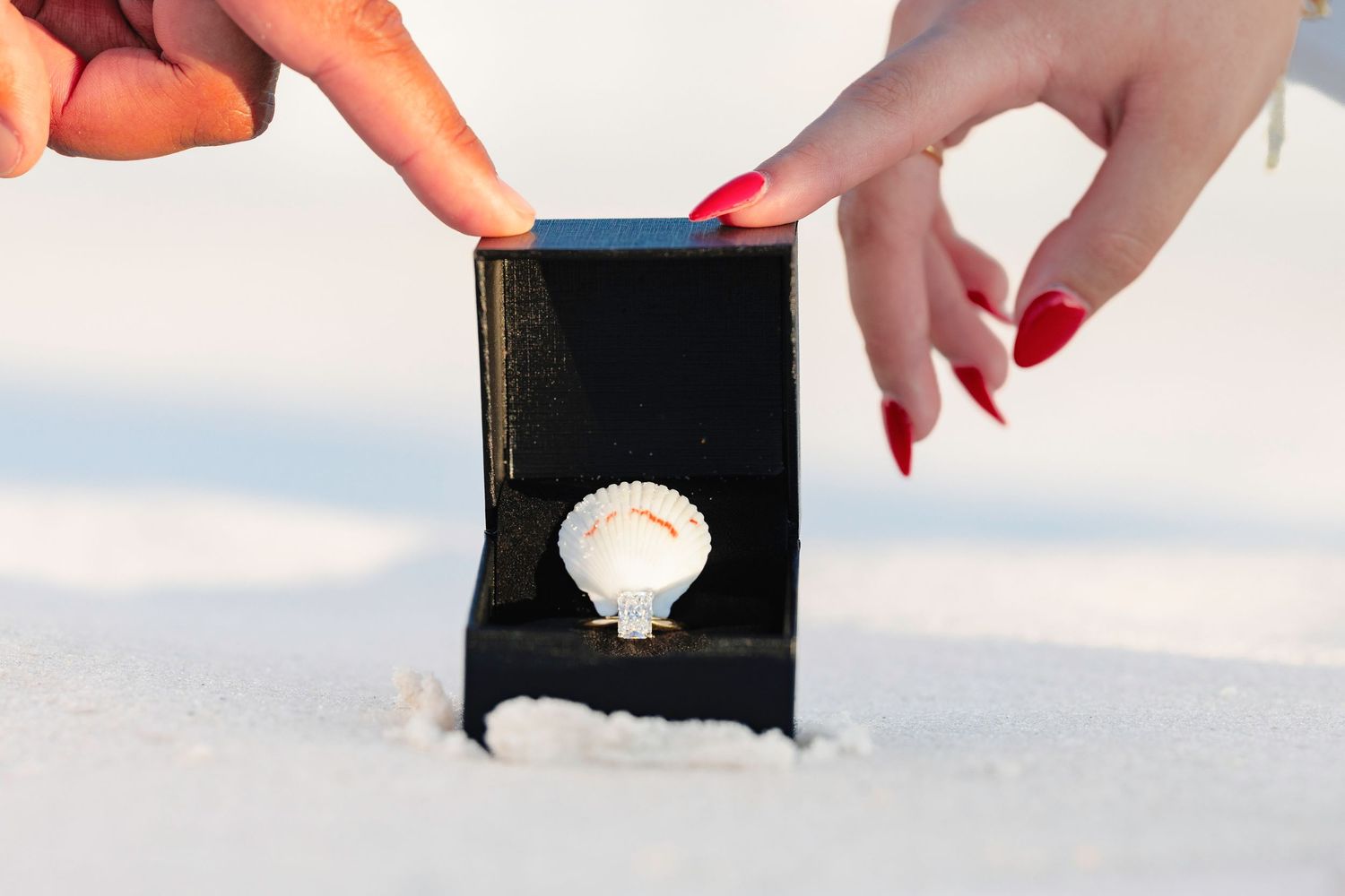 Hands opening a black jewelry box with a seashell ring on white sand beach.